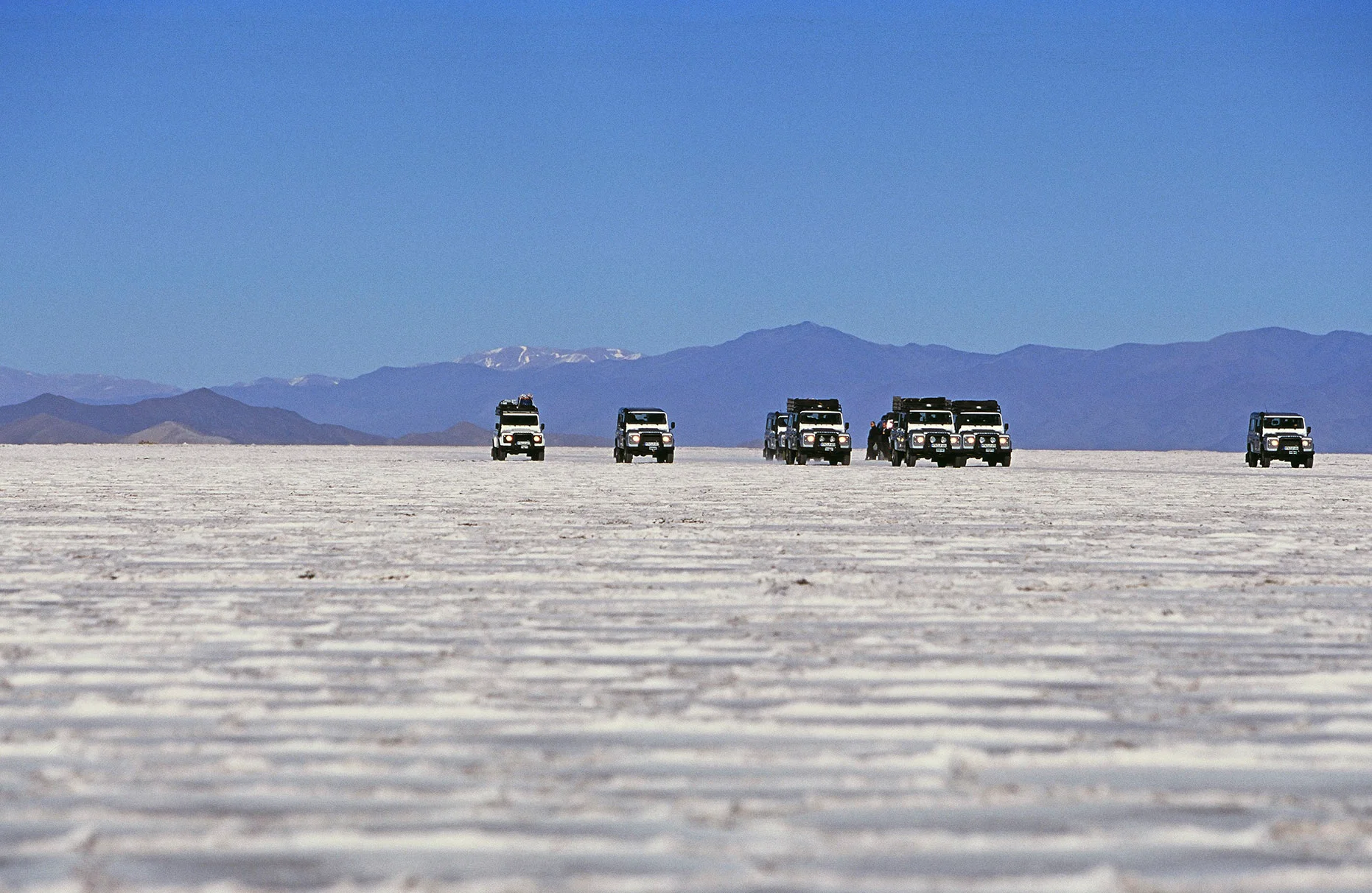 Eine Geländewagen Kolonne fährt über einen Salzsee mit Berglandschaft im Hintergrund in Argentinien während einer Land Rover Experience Tour; aufgenommen für Mens Health Magazin mit FarbDia-Film.