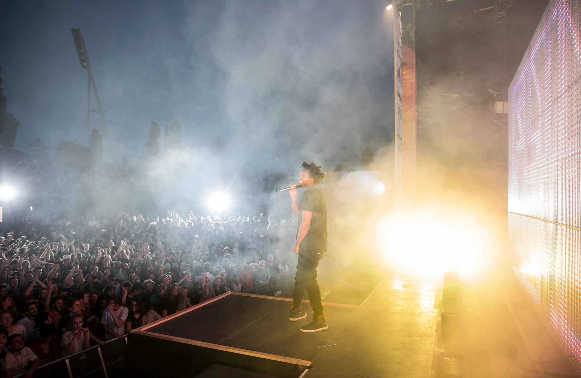 Der Künstler Flying Lotus steht und singt auf der Bühne von Red Bull Music Academy Stage im Mauerpark in Berlin. Im Hintergrund ist der Mauerpark am Abend zu sehen mit viel Nebel und inszeniertem Gegenlicht. Ein zahlenmässig grosses Publikum feiert und jubelt zur Musik.