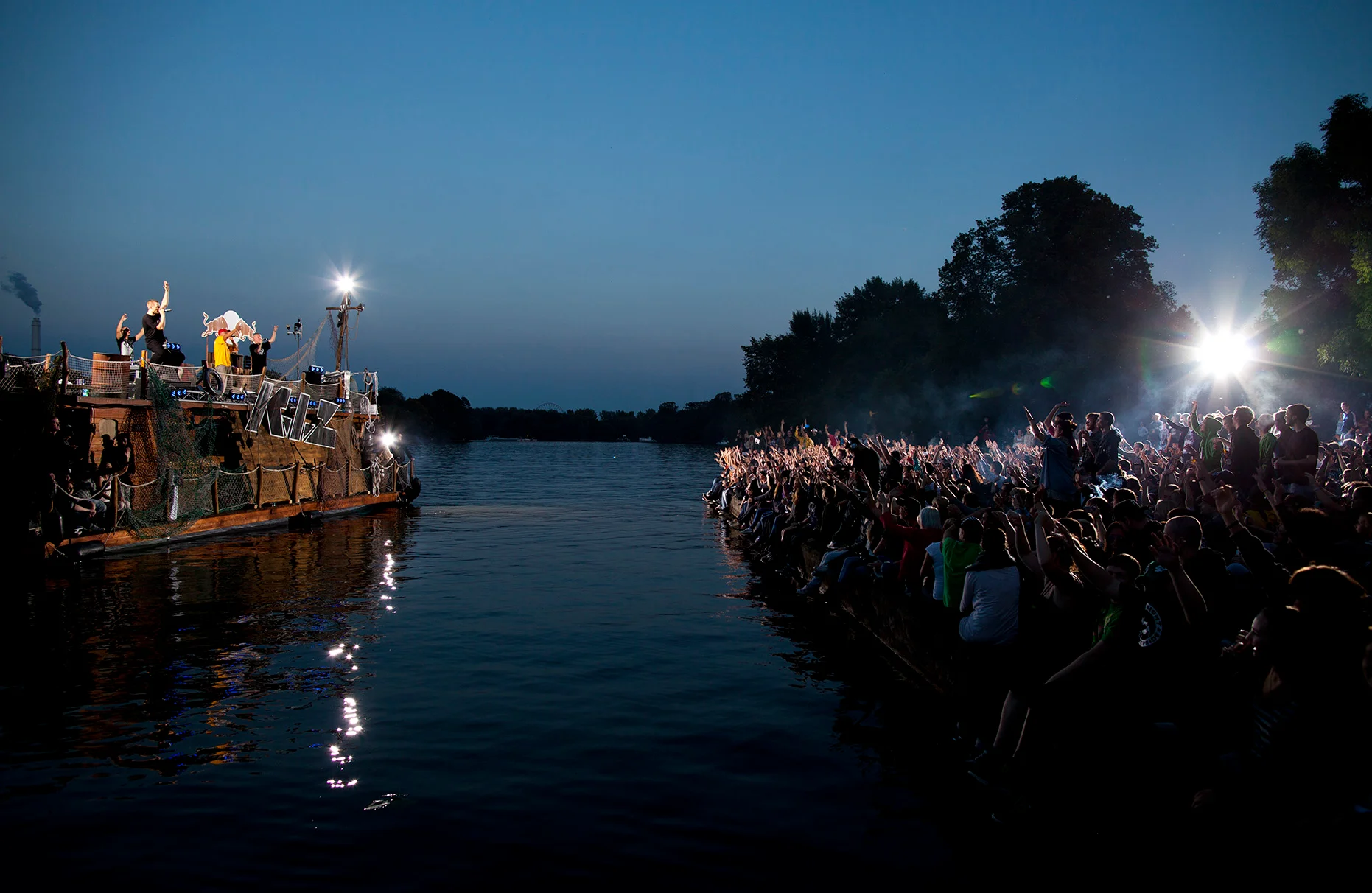 Die deutesche Band K.I.Z singt und spielt auf einem Guerilla-ähnlichem Floss aus Brettern. Das Floss schwimmt auf der Spree am Treptower Park am Abend. Auf der gegenüberliegenden Seite feiern und jubeln die Fans dazu und werden mit inszeniertem Gegenlicht beleuchtet. Die Veranstaltung wurde von Red Bull durchgeführt.