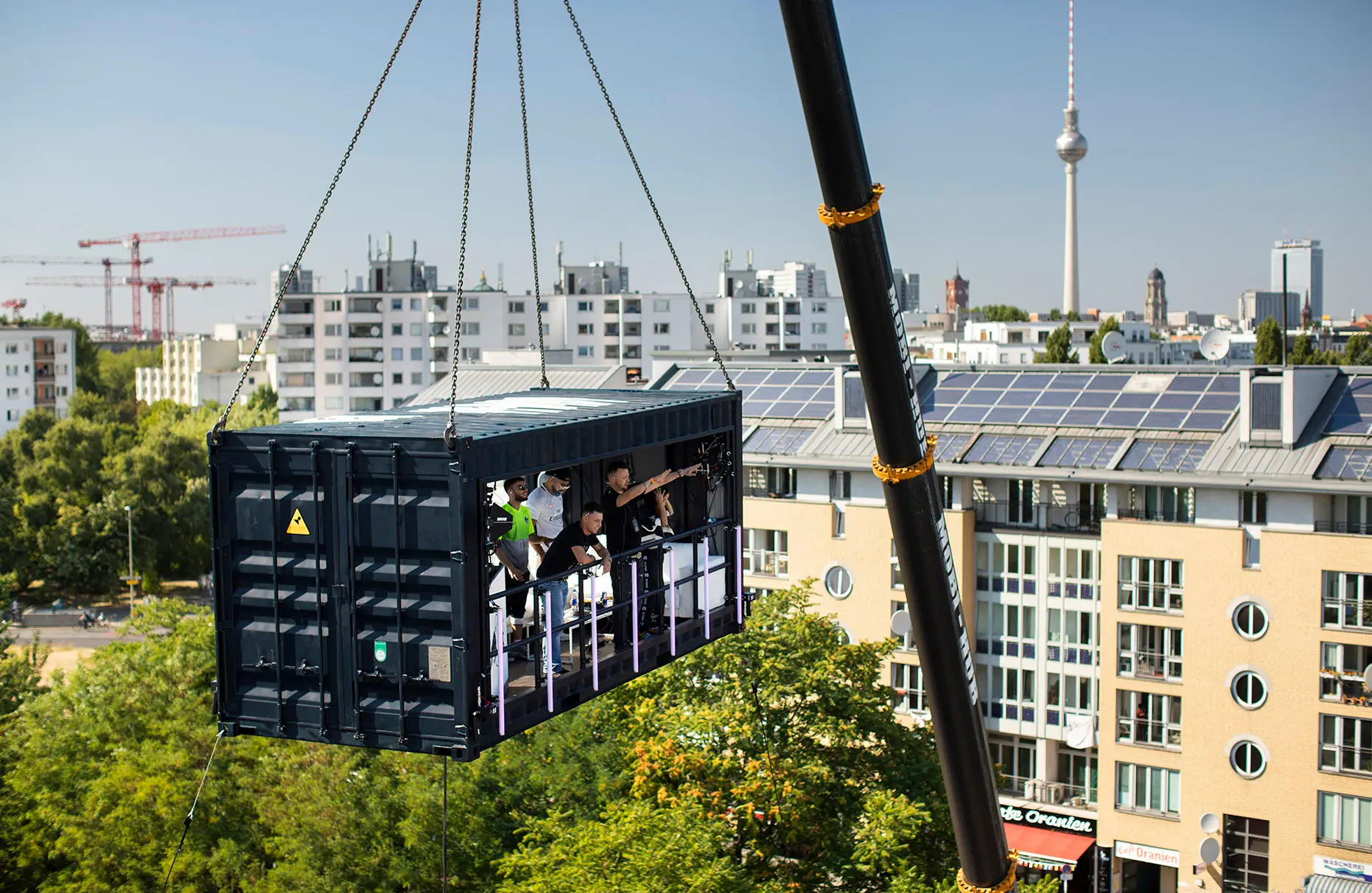 Die deutsche Band KMN-GANG singt und spielt in einem umgebauten Container, der an einem Schwerlastkran hängt. Seinerzeit war dies das höchstschwebende Konzert überhaupt Die Veranstaltung wurde von Red Bull durchgeführt. Im Hintergrund sieht man eine helle, eher moderne Wohnhauskulisse mit Berliner Fernshturm.