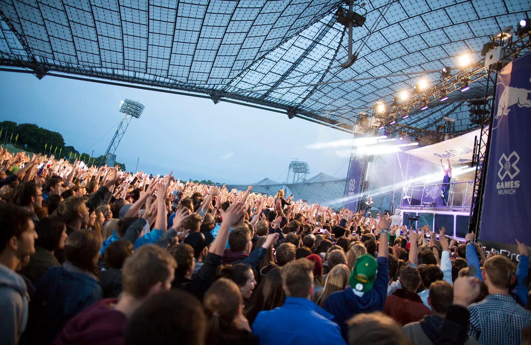 Die Küntlser Maklemore and Ryan Lewis spielen auf der Bühne des Münchner Olympiastadions. Im Hintergrund sind Teile des Olympiastadions am Abend zu sehen und im Vordergrund tobt das Publikum. Das Konzert wurde von Red Bull veranstaltet.