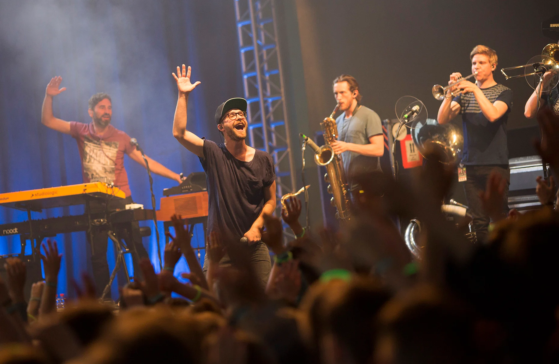 Der Sänger Mark Forster singt auf der Bühne eines AOK Schulkonzertes. Mit auf der Bühne stehen Trompete spielende Musiker. Im Vordergrund jubelt unscharf das Publikum.