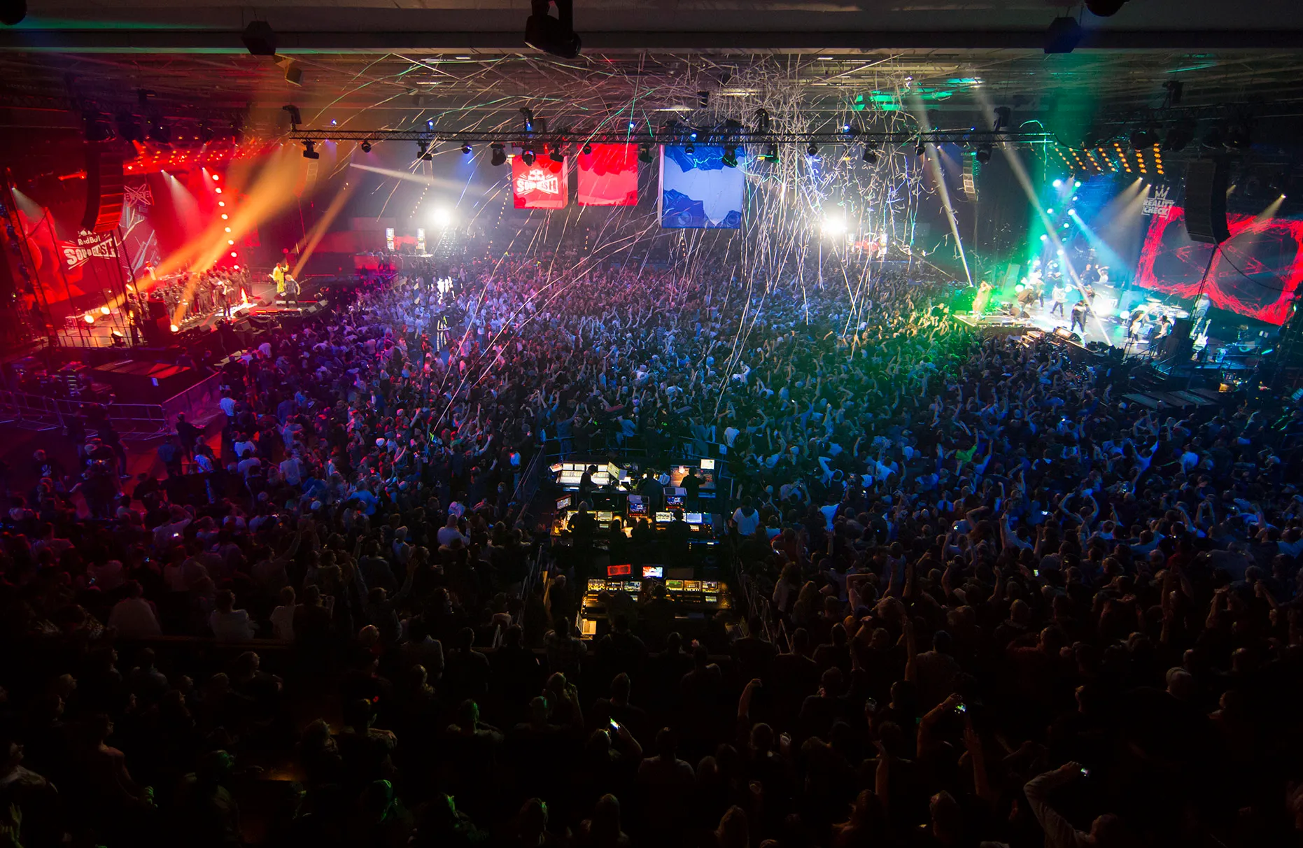 Eine Übersichtsaufnahme des Red Bull sound clash in Hamburg. Es spielen und singen CRACK IGNAZ, LGOONY, SOUFIAN vom Team New Level gegen SAMY DELUXE, EKO FRESH, AFROB vom Team Reality Check. Beide spielenden Bühnen sind zu sehen, dazwischen das tobende Publikum mit viel farbiger Bühnenbeleuchtung. In der Mitte des Saales regent es Luftschlangen.