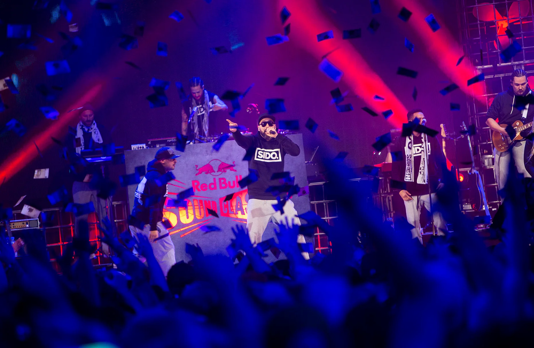 Der Künstler Sido singt auf der Bühne eines Red Bull sound clash in Essen. Sein Rival ist dabei der Künstler Haftbefehl. Im Vordergrund jubelt unscharf das Publikum mit in die Luft gestreckten Armen und es regnet Geldscheine.