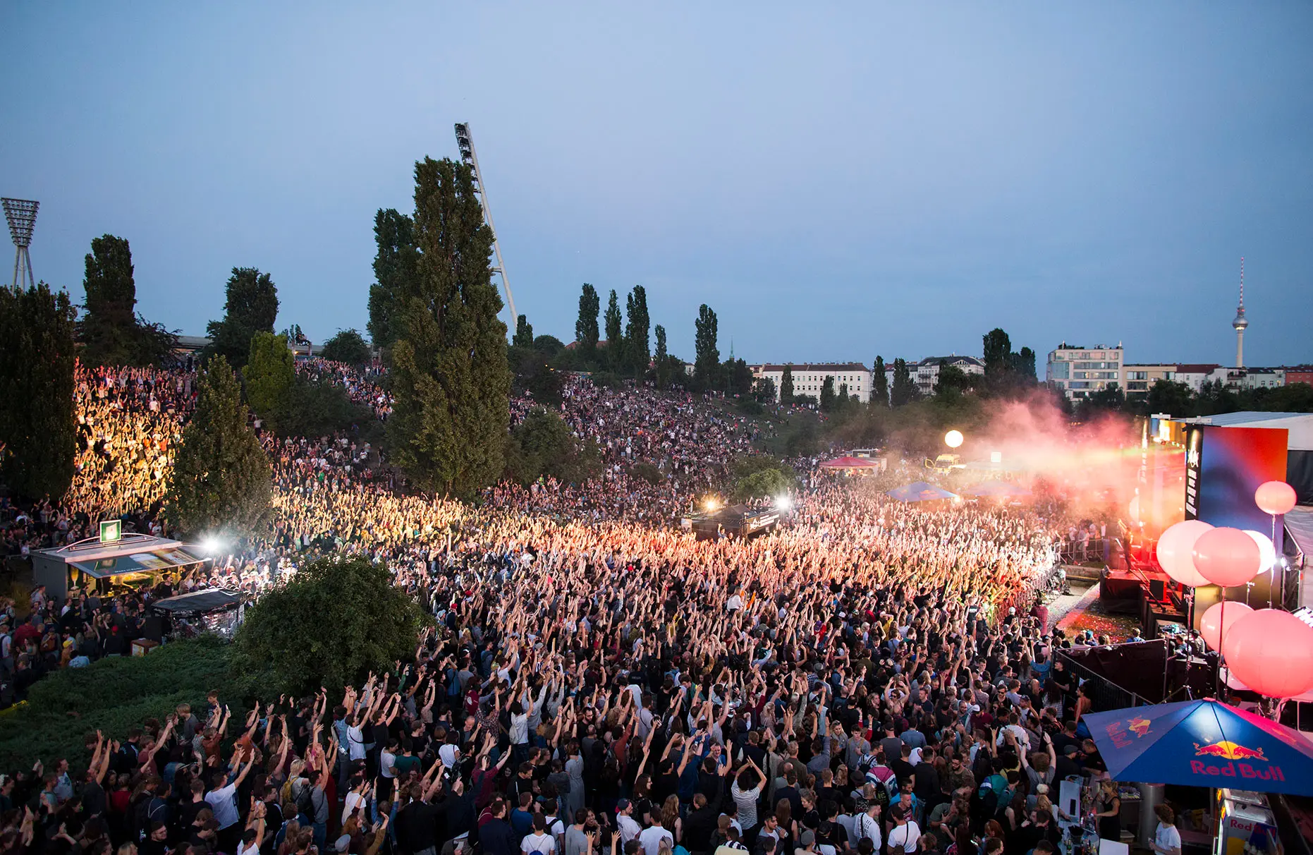 Von einem Steiger bzw. Hebebühne aus fotografiert ist der Mauerpark während eines Red Bull Music Academy Konzertes am Abend zu sehen. Der Park ist fast vollständig mit feiernden Menschen gefüllt. Das Bühnenlicht und Blitzlicht sorgt für viel farbige Beleuchtung im Bereich der feiernden Masse. Am Horizont der Prenzlauer Berg und der Fernsehturm.
