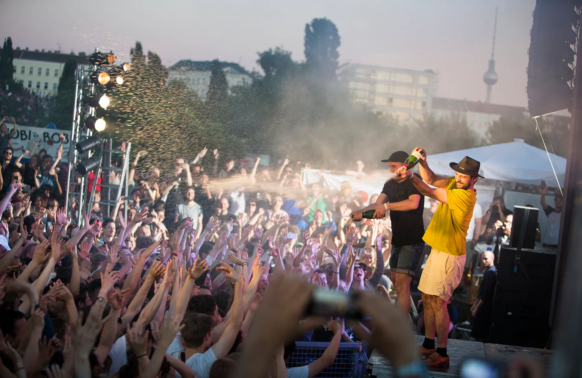 Das Künstlerduo modeselector spielt während eines Red Bull Music Academy Konzertes. Die Bühne steht im Mauerpark in Berlin. Die Beiden bespritzen das tobende Publikum mit Sekt aus geschüttelten Flaschen. Das Bühnenlicht und Blitzlicht sorgt für farbige Beleuchtung. Am Horizont unscharf Häuser des Prenzlauer Berges und der Fernsehturm.