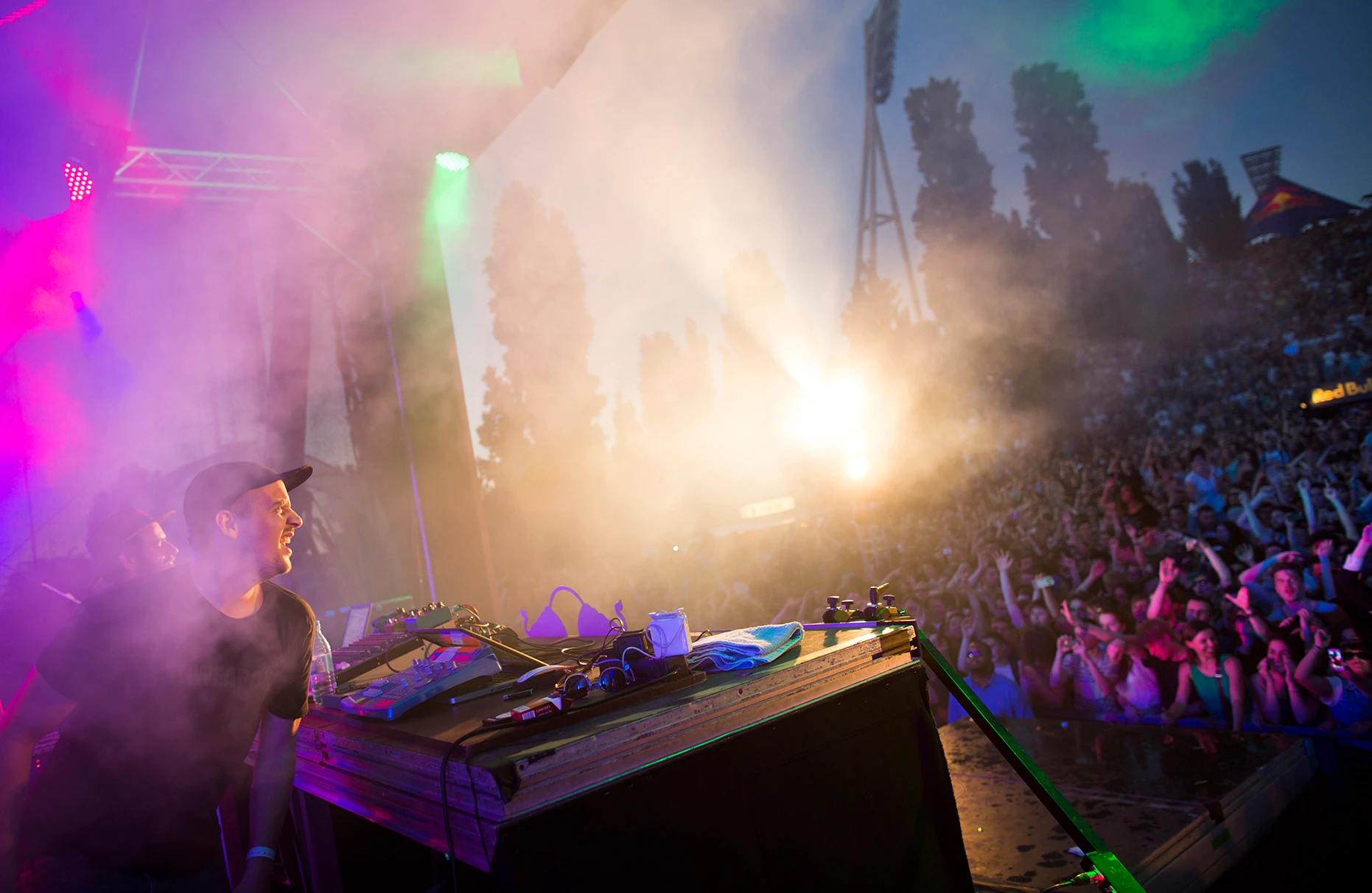 Das Künstlerduo modeselector spielt ausgelassen an den turntables auf der Bühne der Red Bull Music Academy im Mauerpark in Berlin. Im Hintergrund ist das tobende Publikum in farbiger Beleuchtung, Nebel und künstlichem Gegenlicht zu sehen. Am Horizont unscharf die hohen Pappeln des Mauerparks am Abend.
