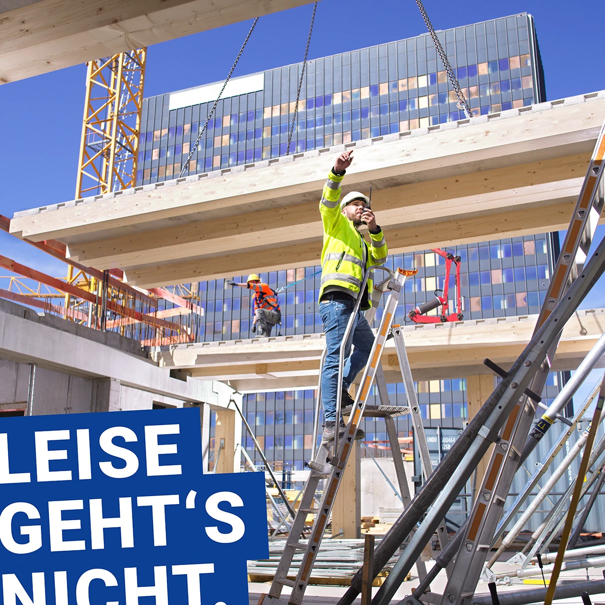Ein Arbeiter kontrolliert und unterstützt die an einem Kran hängende Holtzbalken- Last während der Beförderung auf der oberen Etage eines nachhaltigen Neubaus. Im Hintergrund ein verglastes Geschäftsgebäude und blauer Himmel. Produziert für eine Werbekampagne des Hauptverbands der dteutschen Bauindustrie e.V. mit Hasselbald 501c CFVii50v.