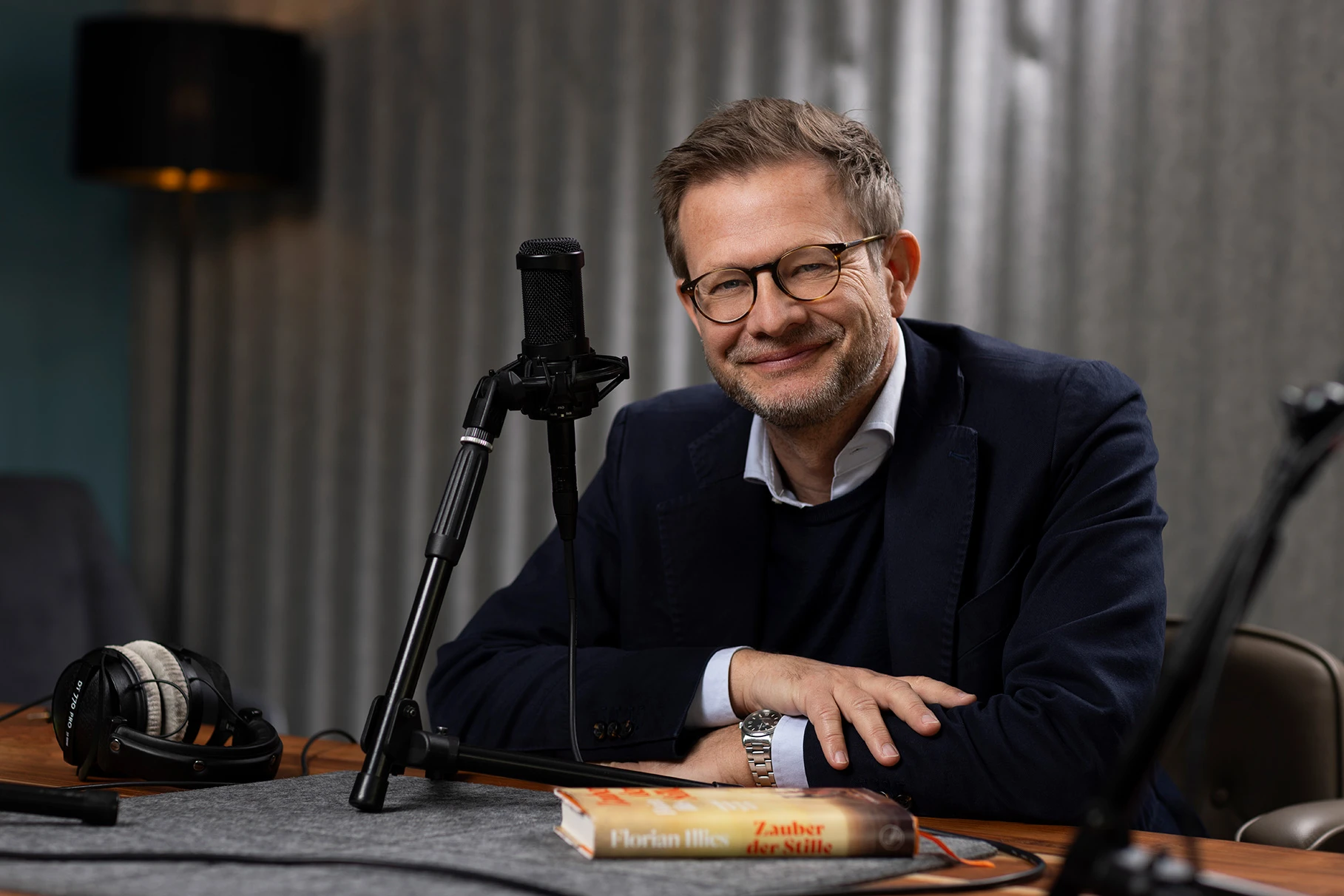 Der Autor Florian Illies sitzt an einem Tisch mit Tontechnik in einem Aufnahmestudio für podcast's und schaut lächelnd in die Kamera. Produziert für Visit Berlin Tourismus & Kongress GmbH.