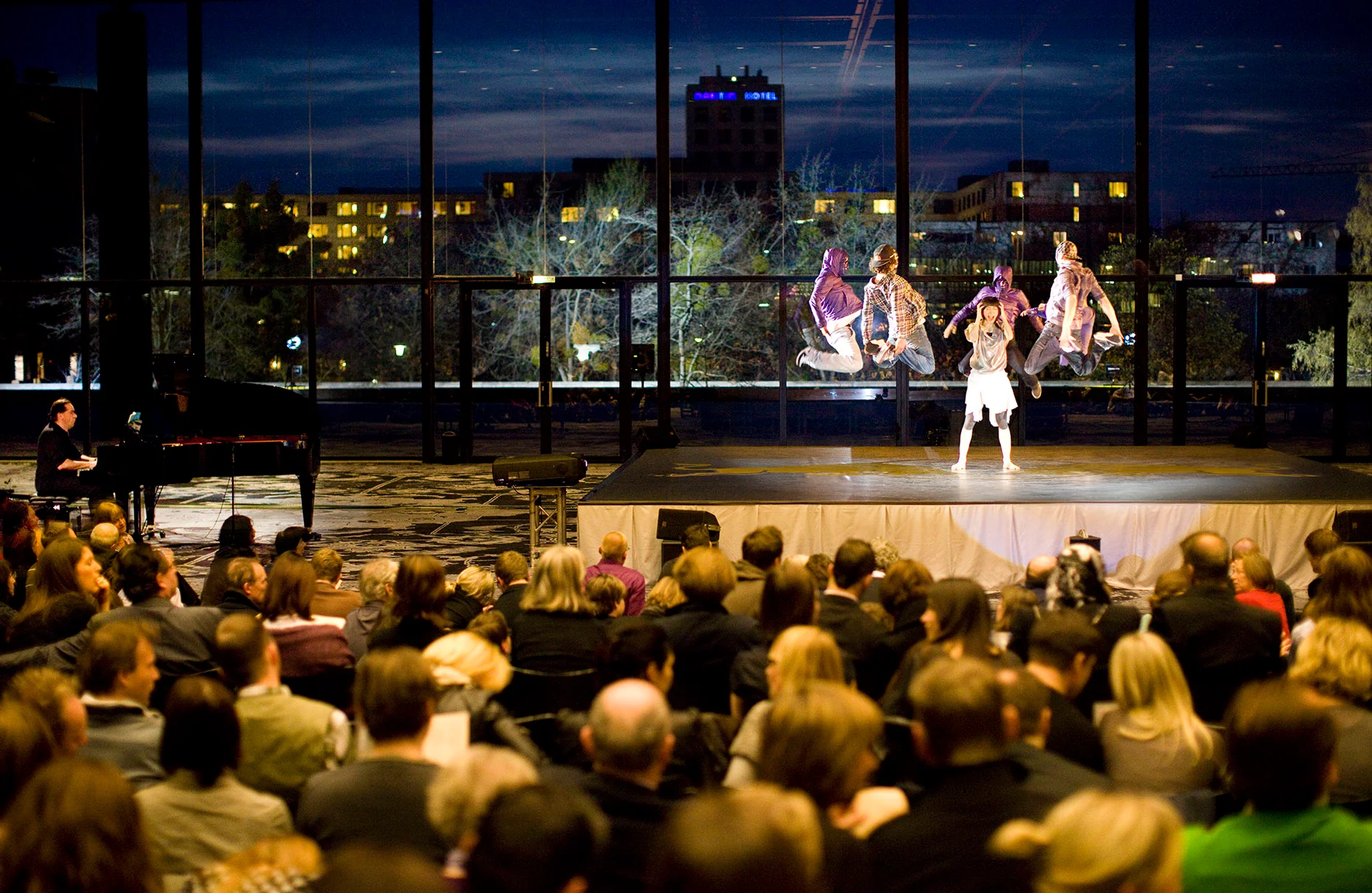 Die breakdance crew Flying Steps in Aktion während der Premiere Vorführung von Flying Bach in der Nationalgalerie in Berlin vor Publikum in den Sitzen. Aufnahme leicht von oben mit weitwinkliger Sicht auf Räumlichkeit und Stadtkulisse durch die Fensterfassade bei abendlichem Licht. Prodziert für Red Bull.
