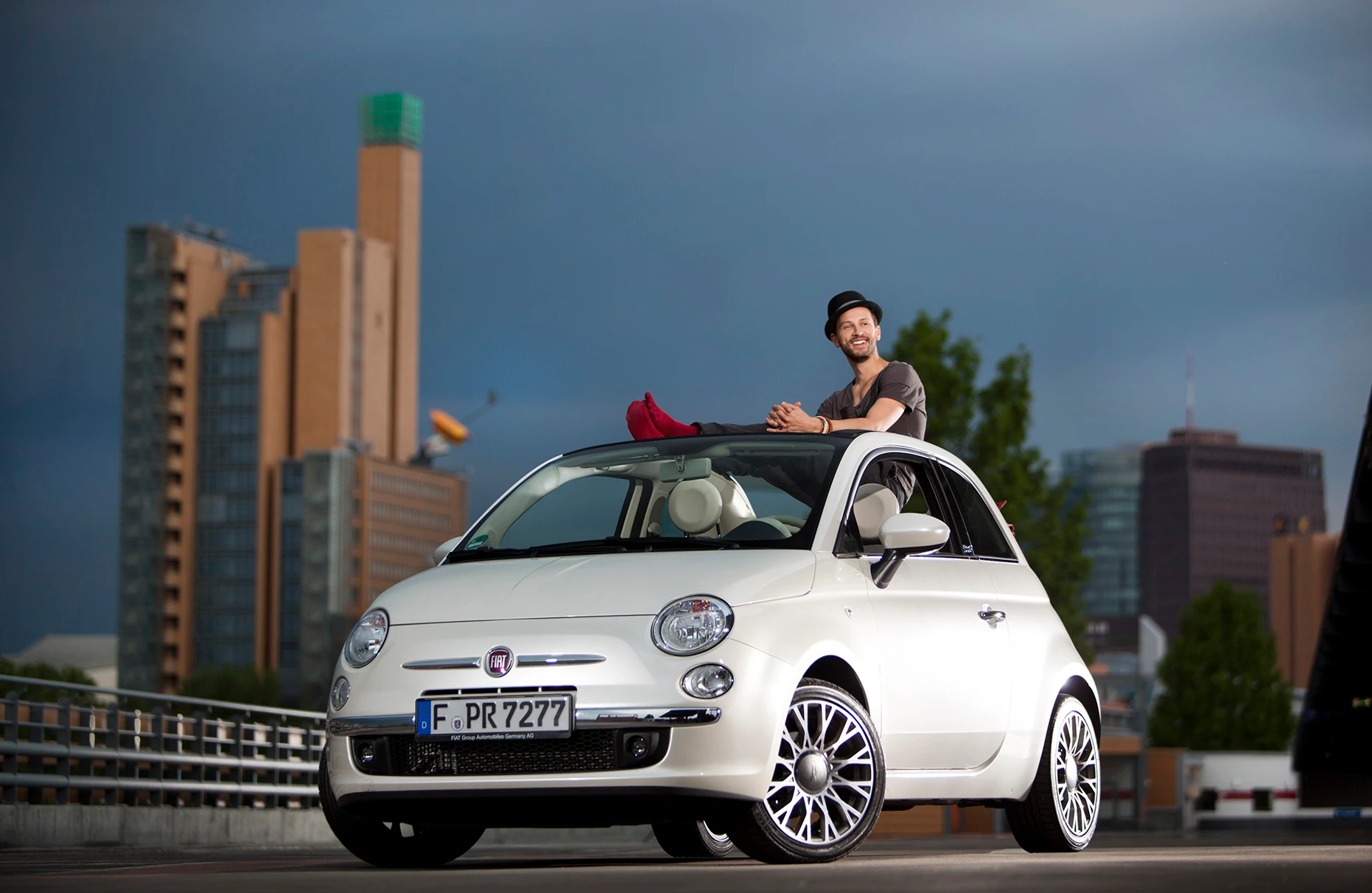 Der Schauspieler Franz Dinda sitzt auf einem Fiat 500 Cabrio auf dem Dach eines Parkhauses und lächelt. Im Hintergrund ist der Potsdamer Platz von Berlin bei abendlich blauem Himmel zu sehen. Produziert im Rahmen einer Fiat PR Kampagne für Schröder & Schömbs PR.