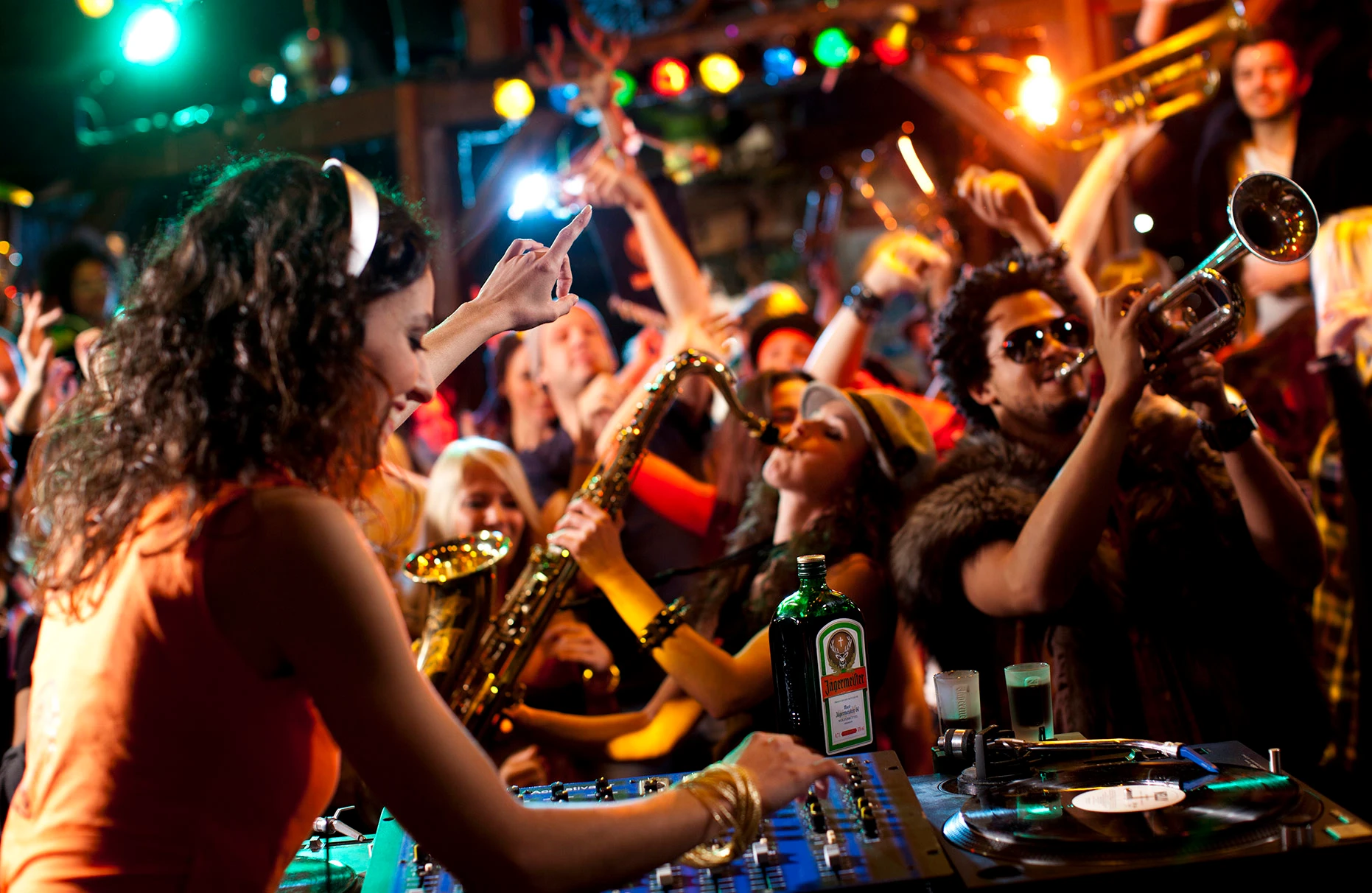 Die kunterbunt gekleideten Mitglieder der Jägermeister Blasskapelle spielen euphorisch in einer Disco. Im Vordergrund eine Djane mit Jägermeisterflasche an den turntables. Bunte Disco-Beleuchtung. Produziert für eine Jägermeister PR- Kampagne von White Rabbit PR.