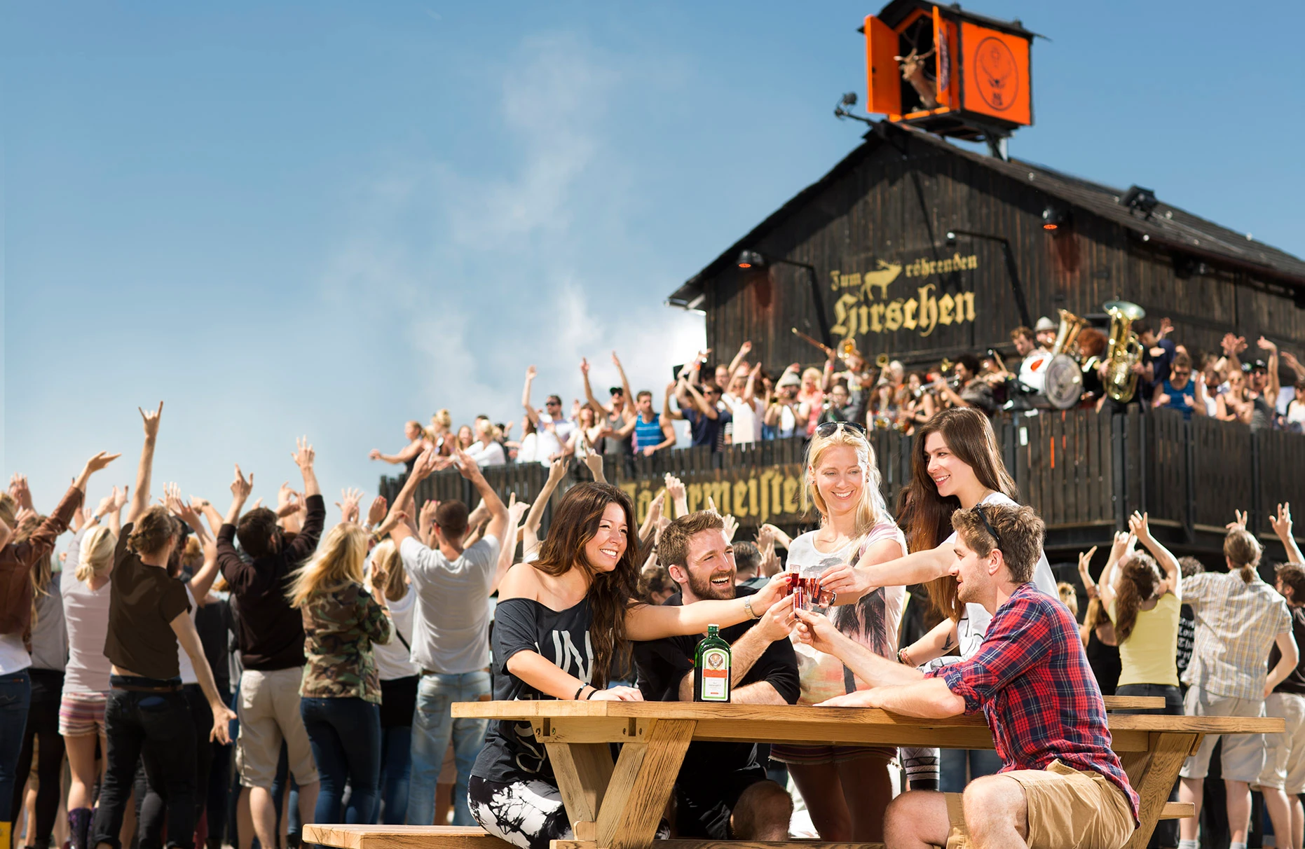 Die kunterbunt gekleideten Mitglieder der Jägermeister Blasskapelle spielen euphorisch auf der Balkon Terasse einer Holzhütte auf einem Musik-Festival. Im Vordergrund feiernde Gäste. Ganz vorn im Fokus ein Holztisch mit Jägermeister anstossenden , fröhlichen Gästen. Produziert für eine Jägermeister PR- Kampagne von White Rabbit PR.