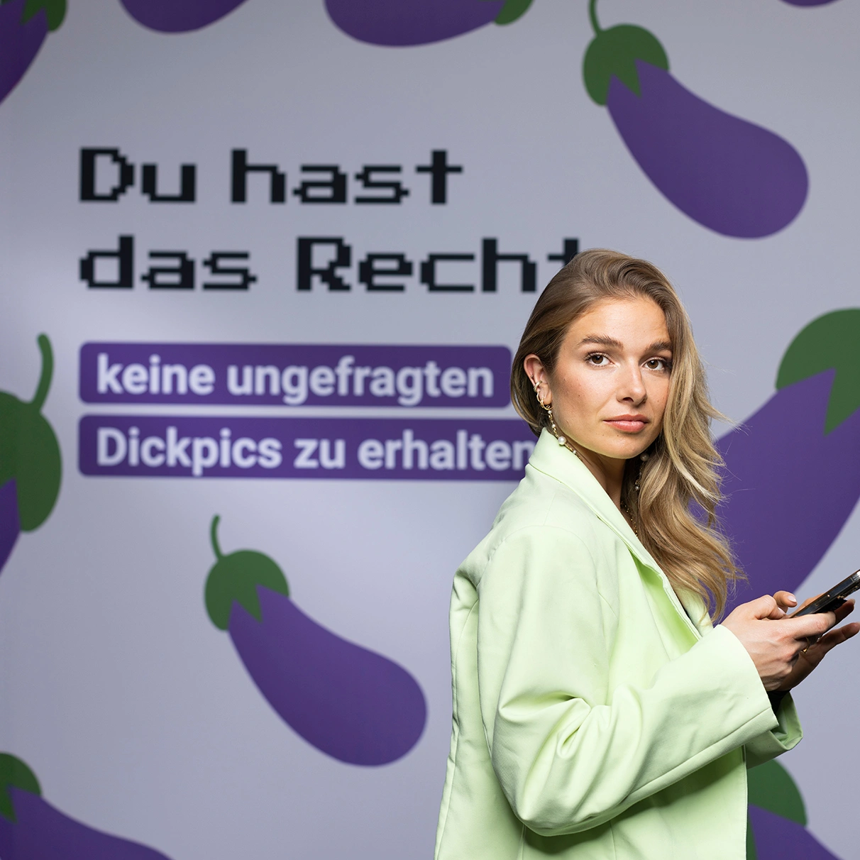 Die Moderatorein Lola Weipert steht in modischem outfit und Handy mit Blick in die Kamera vor einer Leinwand mit einer Grafik und dem Spruch: Ich habe das Recht, keine ungefragten dickpics zu erhalten. Prodziert für eine Kampagne der NGO Hate Aid.