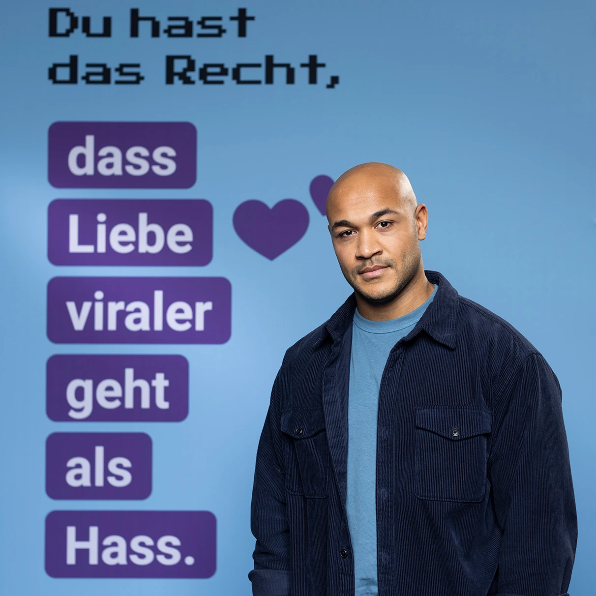 Der Schauspieler Malick Bauer steht mit Blick in die Kamera vor einer Leinwand mit einer Grafik und dem Spruch: Du hast das Recht, dass Liebe viraler geht als Hass. Prodziert für eine Kampagne der NGO Hate Aid.