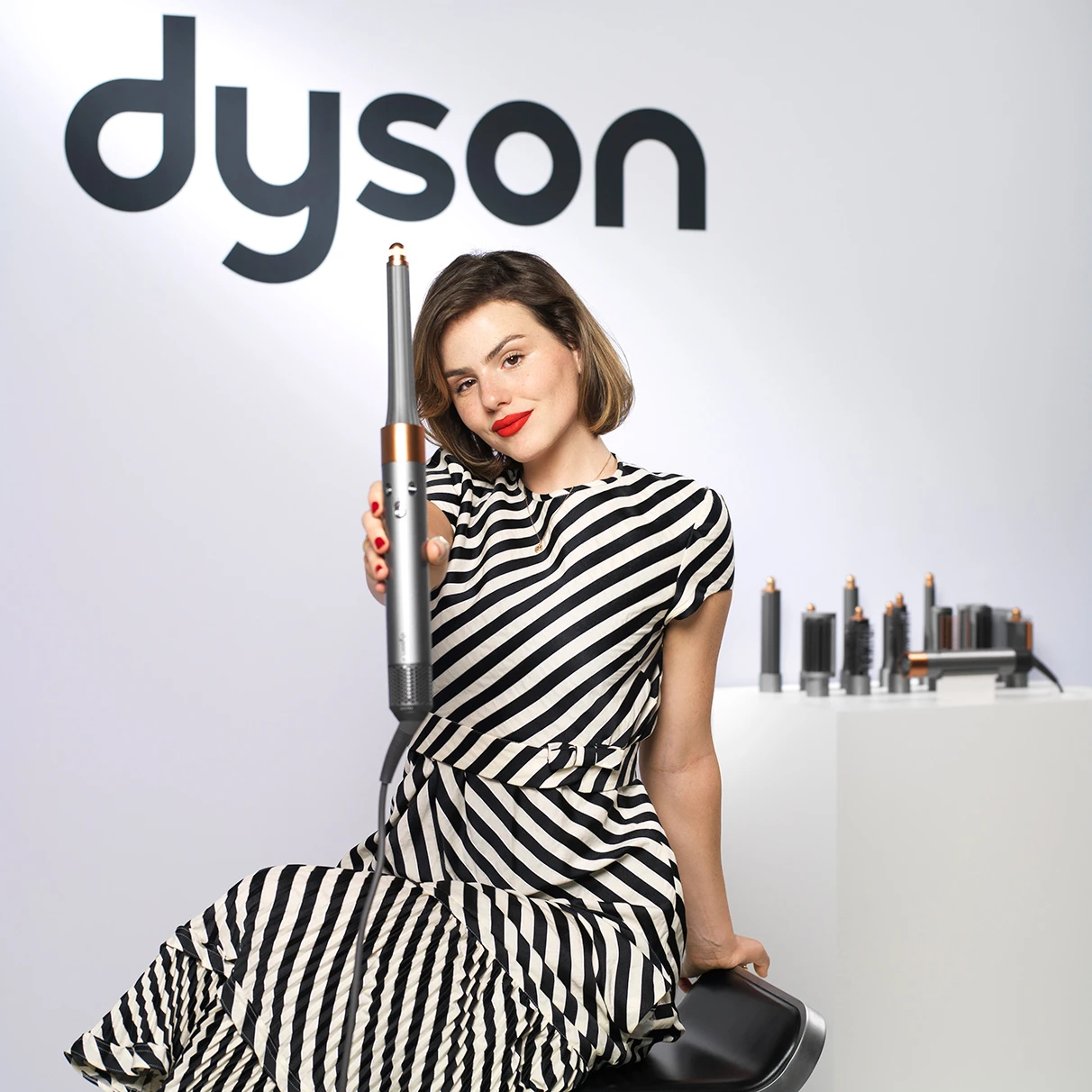Die Schauspielerin Ruby O. Fee sitzt auf Barhocker mit Dyson Lockenstab vor weisser Wand mit Dyson-Logo und läcjelt in die Kamera. Produziert anlässlich einer Dyson PR Kampagne in Hamburg mit Hasselblad 501c CFV16