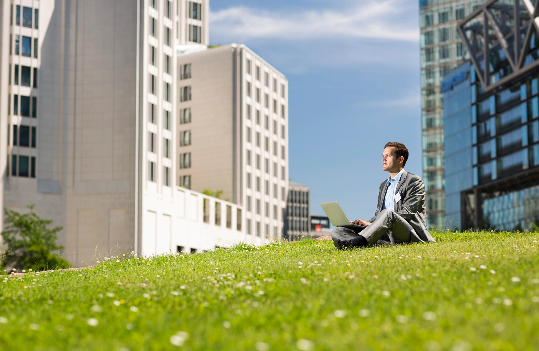 Ein junger business Mann sitzt mit laptop auf einer grünen Wiese am Potsdamer Platz und schaut nachdenkend. Blauer Himmel. Prodziert für eine Kampagne von Visit Berlin Tourismus & Kongress GmbH.