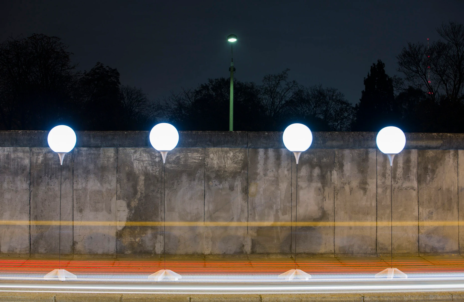 Zu sehen ein Stück Berliner Mauer in der Bernauer Strasse. Frontalansicht mit Lichtstreifen von vorbeifahrenden Autos. Anlässlich der 25 Jahre Mauerfall Feier stehen 4 Licht- Ballons, die zu einer langen Kette entlang der ehehmaligen Mauer gehören. Produziert für Visit Berlin Tourismus & Kongress GmbH