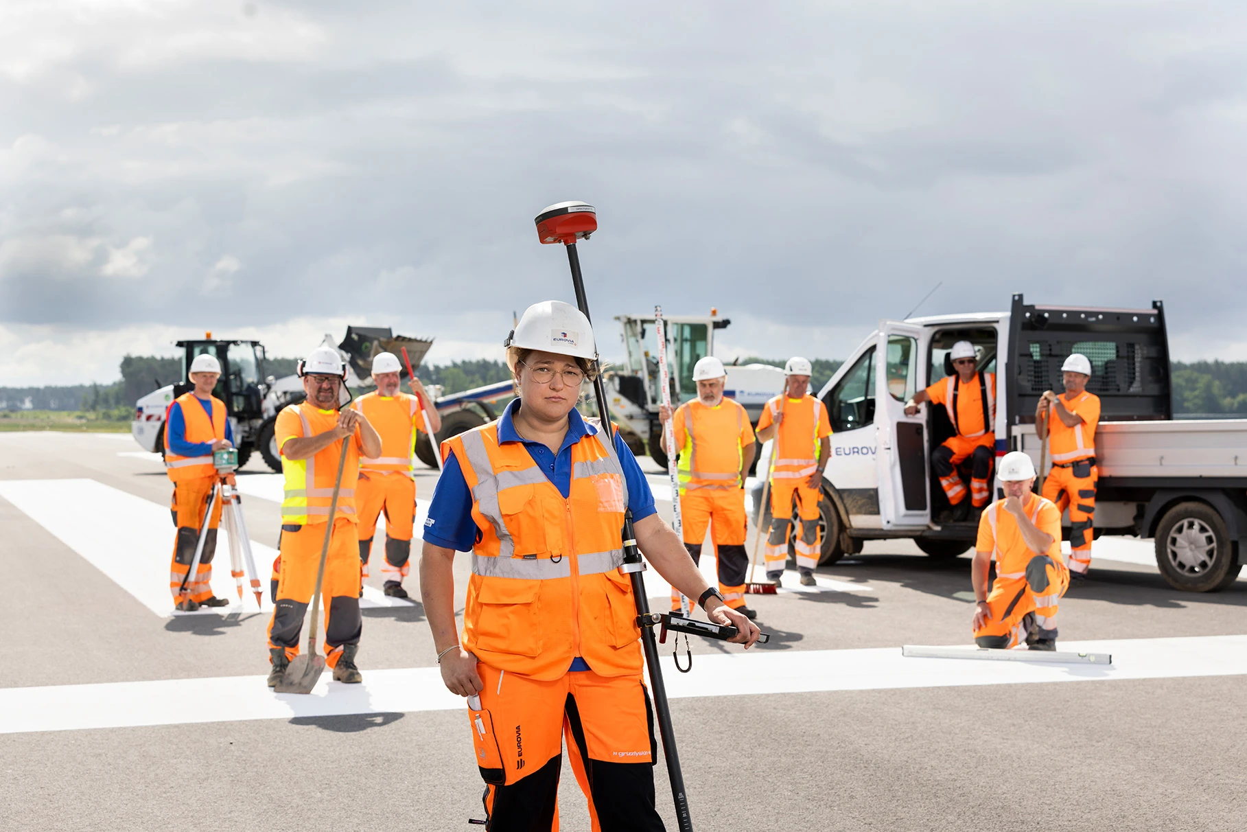 Ein Asphalt Strassenbau- Team posiert mit Radlader und ähnlichen Maschinen- Fahrzeugen auf einer Rollbahn eines kleinen Flughafens bei Sonnenschein. Im Vordergrund eine weibliche Arbeiterin in PSA. Produziert für Eurovia Vinci.