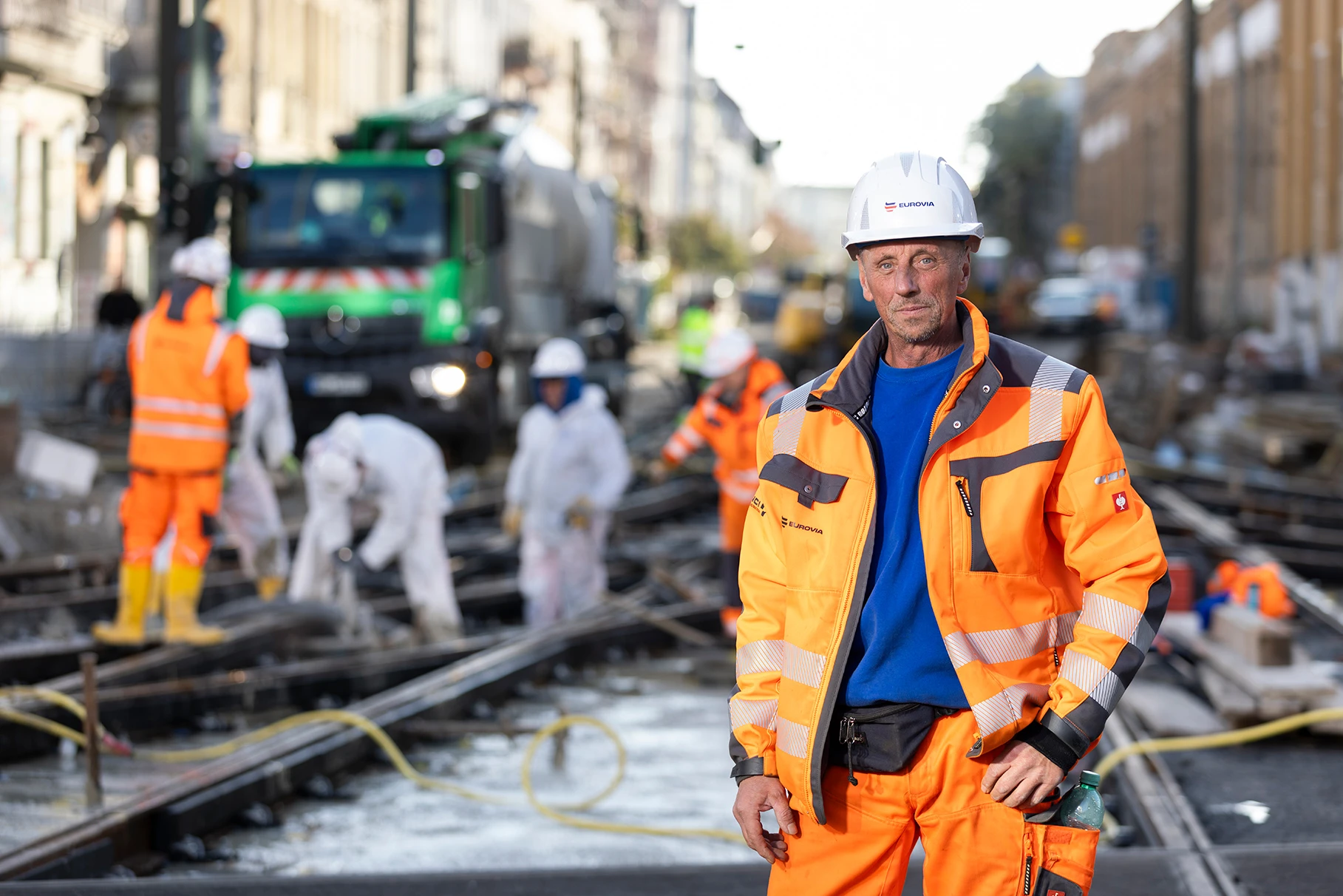 Ein Strassenbau- Arbeiter in PSA posiert auf einer Baustelle in Berlin Schöneweide. Im Hintergrund die Strassenschlucht mit seinen Kollegen, die eine der kompexesten Tram- Gleisanlagen Deutschlands frisch mit Asphalt füllen. Produziert für Eurovia Vinci.