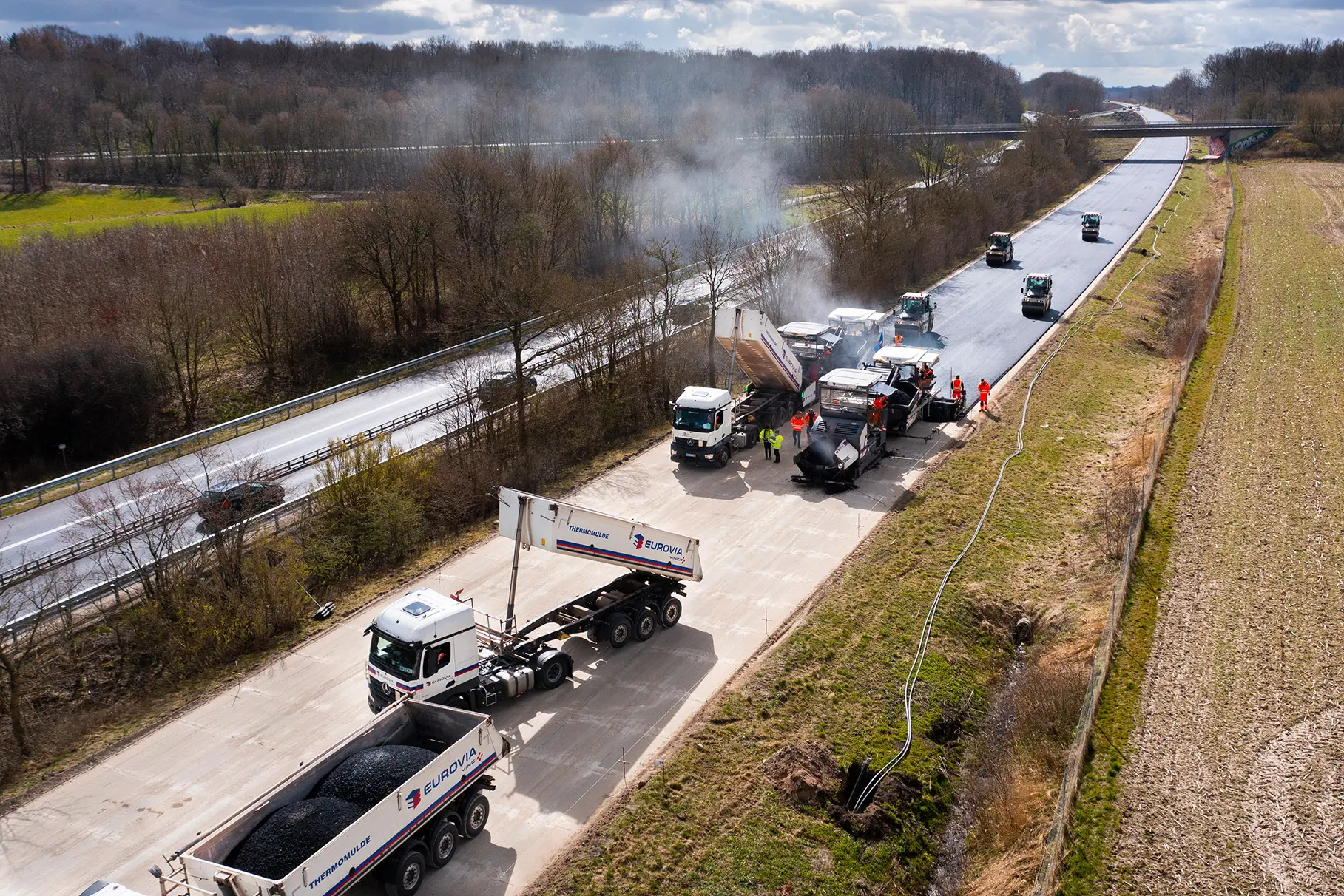 Die A215 Autobahn wird mit zwei Asphalt- Fertigern, Walzen und LKW saniert. Assphalt- Baustelle. Rundherum Natur und bewölkter Himmel. Aufgenommen für EUROVIA Teerbau GmbH NL Hamburg mit einer DJI Drohne.