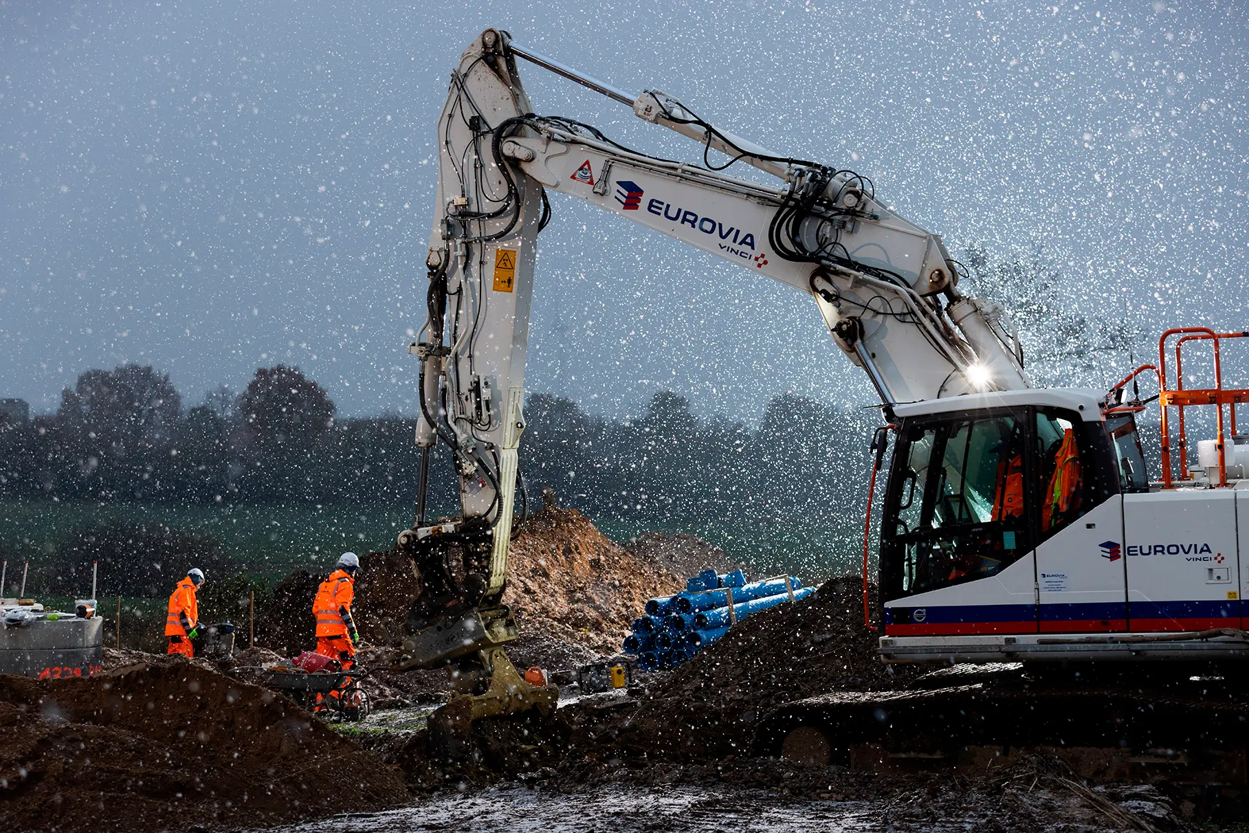 Ein Bagger während Bauarbeiten in einer Tiefbau- Baustelle von Eurovia mit zwei orangefarben gekleideten Arbeitern. B-Plan am frühen morgen mit Natur im Hintergrund. Es fällt Schnee, der sich durch künstliches Blitz- Gegenlicht abhebt. Produziert für Eurovia Vinci.
