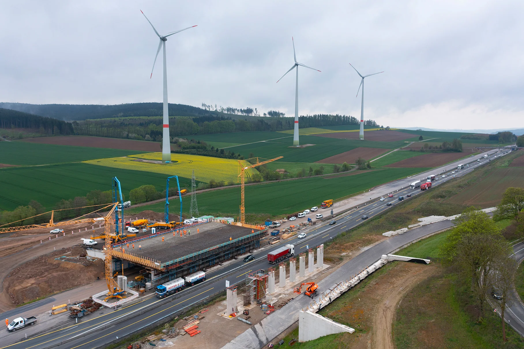 Die Autobahn A7 diagonal durch das Bild laufend in einer Vogelperspektive im Frühjahr mit bedecktem Himmel und Windrädern. Über die Fahrbahn wird gerade bei laufendem Betrieb eine Wildbrücke gebaut. Die Hälfte ist fertig. Produziert für Eurovia Vinci mit DJI Drohne.