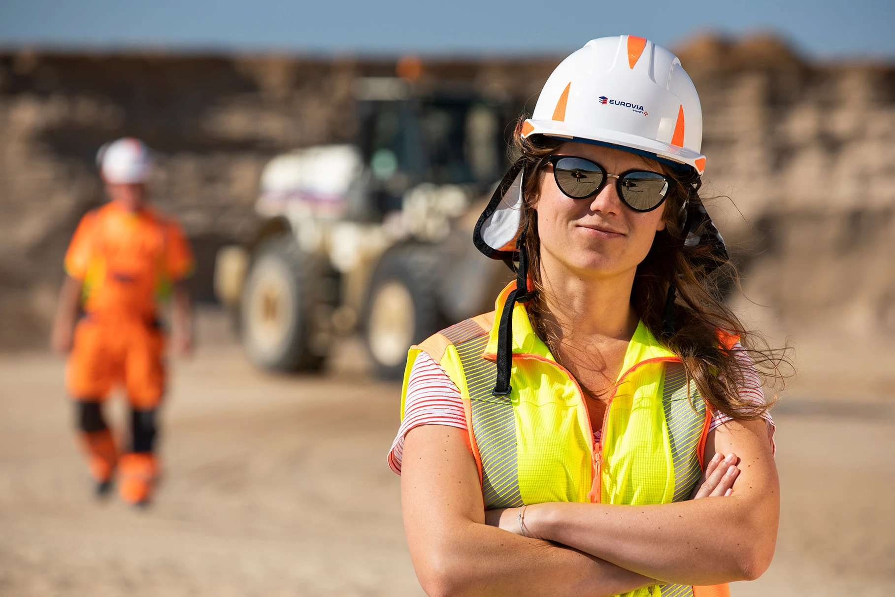 Eine junge Mitarbeiterin in PSA Kleidung und Sonnenbrille steht im Vordergrund bei Sonnenschein. Im Hintergrund ein weiterer Mitarbeiter in PSA und Radlader in einer Kiesgrube. Produziert für Sand und Kies Union GmbH Berlin-Brandenburg, Eurovia Vinci