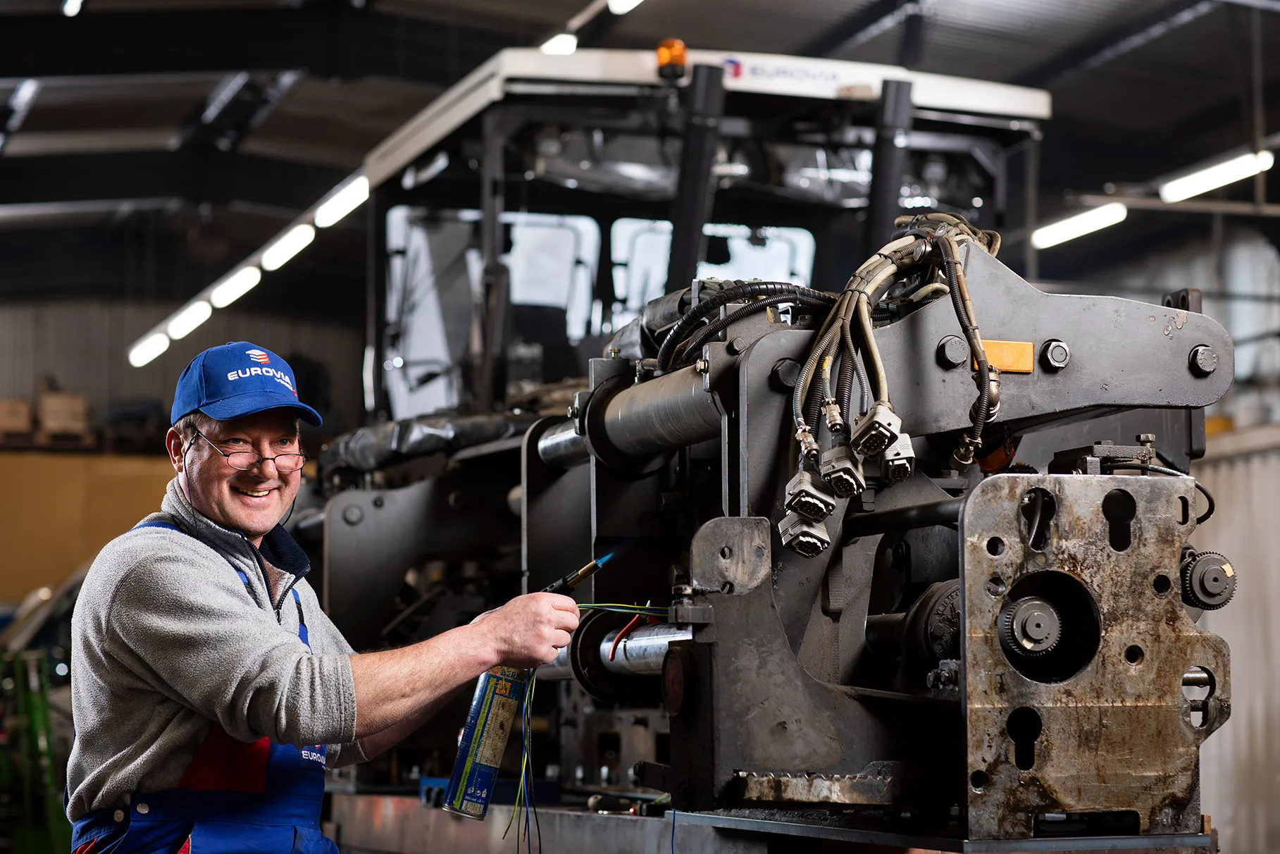 Ein Mechaniker mit Basecap repariert in einer Werkstadt-Halle einen Asphalt- Fertiger und lacht. Produziert für Eurovia Vinci.