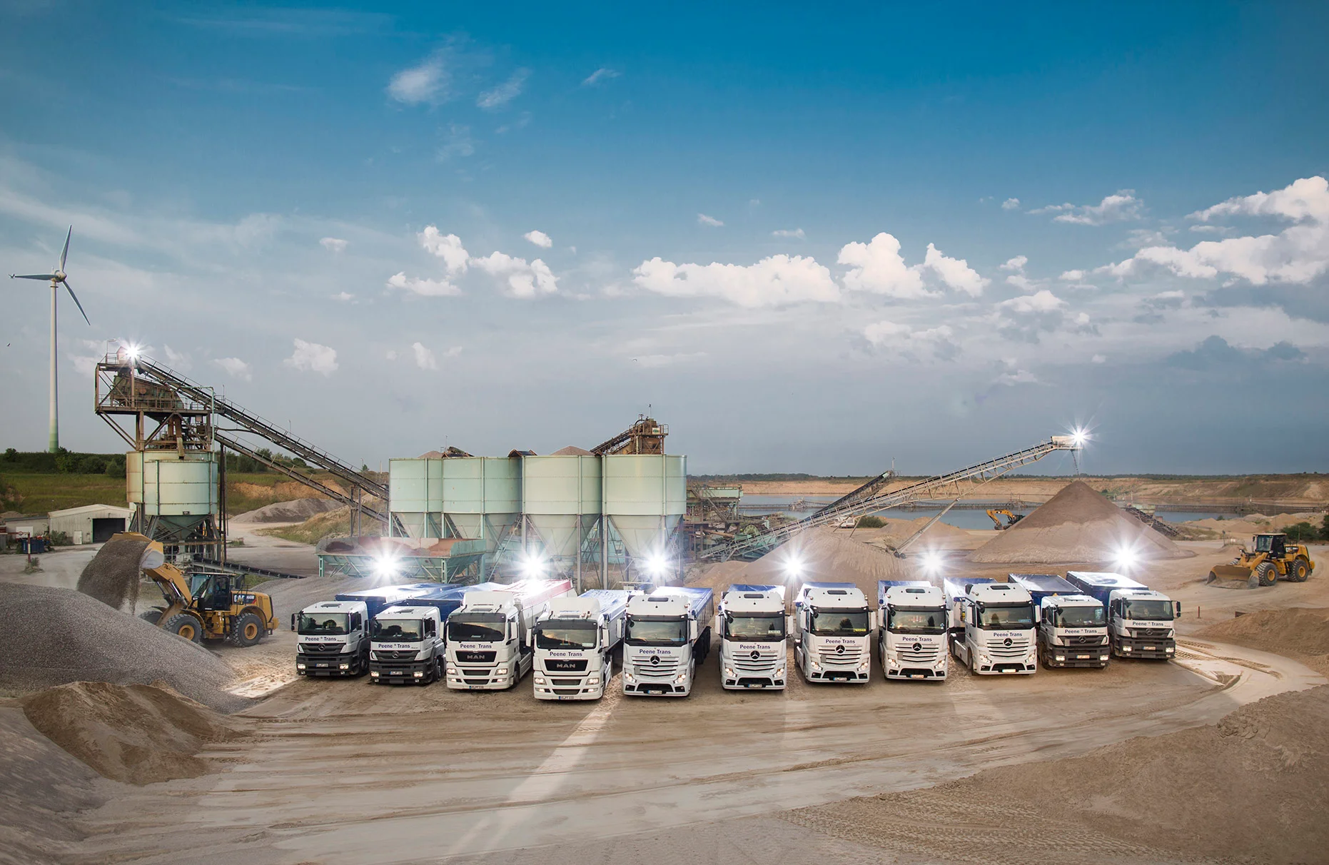 Einige LKW nebeneinander aufgerreiht und mit künstlichem Blitz-Gegenlicht stehen in einer Kiesgrube mit Förderband- Anlagen und Radladern. Blauer Himmel und Windrad am Horizont. Produziert für Peene Trans GmbH.