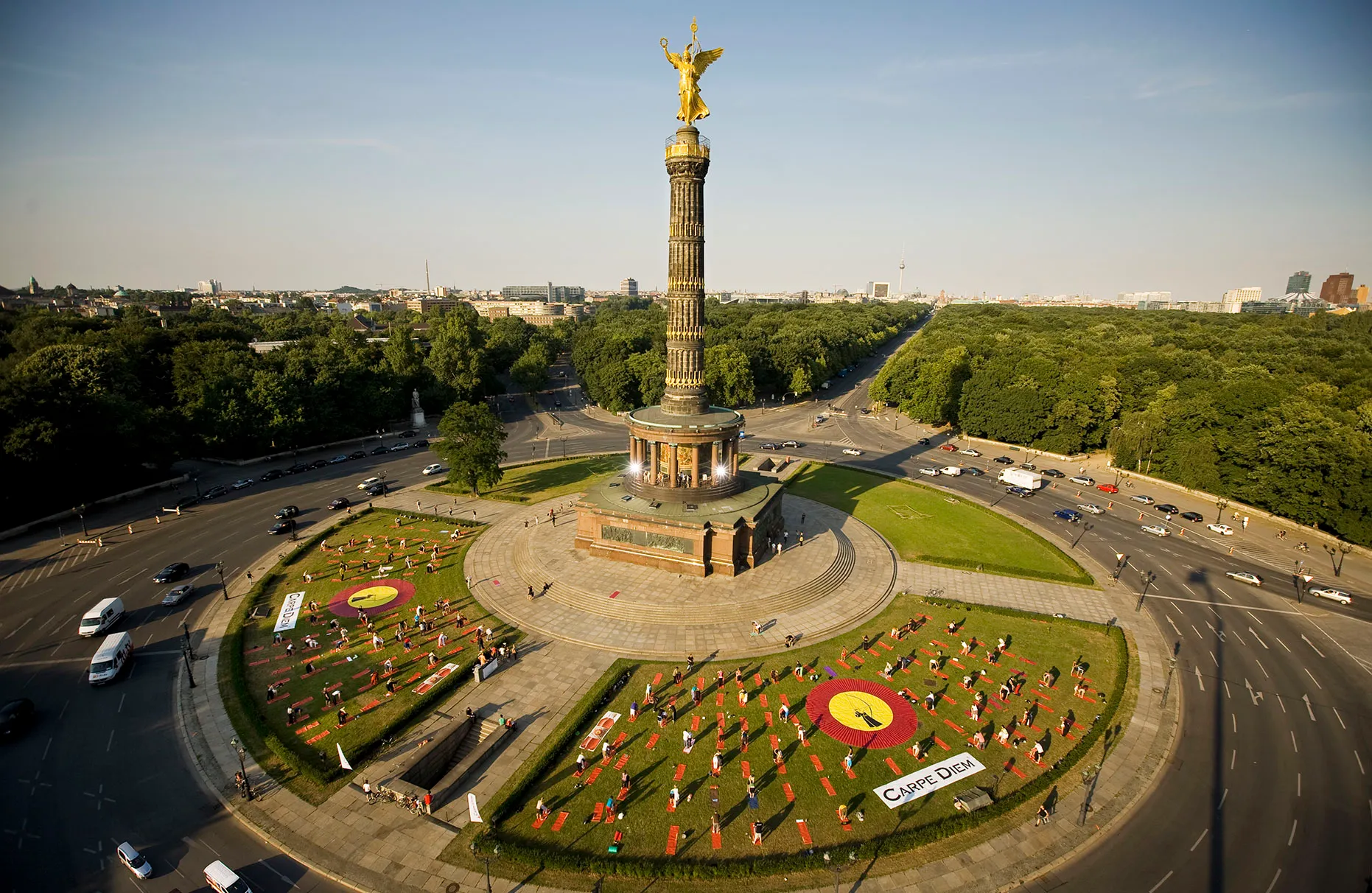 Auf dem Rasen des Kreisverkehr der Siegessäule in Berlin machen Menschen Yoga. Im Hintergrund der Tiergarten bei Sonnenschein im Sommer. Von oben mit Hilfe einer sehr hohen Hebebühne fotografiert für eine PR- Kampagne von Carpe Diem.