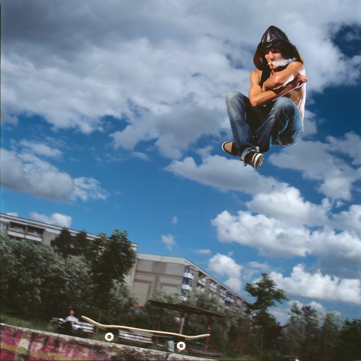 Der skater Rolandi "Hirschi" fliegt in einem skate-Park nach dem Absprung im Schneidersitz durch die Luft. Er trägt nur eine Jeans und Oberkörper frei. Aus seinem Mund pusstet er dabei Zigarettenqualm. Im Hintergrund Plattenbauten und blauer Himmel mit weissen Wolken. Eine Freie Arbeit, aufgenommen mit Hasselblad 501c Farbnegativ-Film.