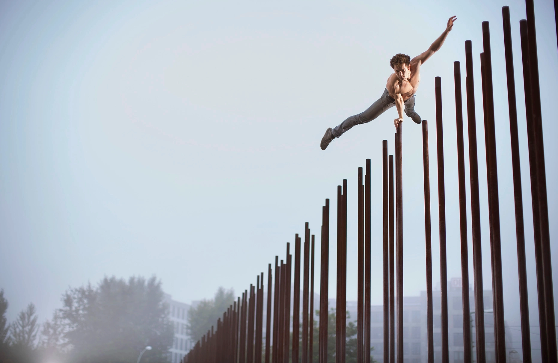 Der Akrobat und Freerunner John Förster posiert mit einer Hand in einer Art Superman- Pose auf den sehr hohen Eisen- Stelen der Gedenkstätte Berliner Mauer in der Bernauer Strasse. In aller frühe fotografiert, noch nebelig und menschenleer. Eine Freie Arbeit.