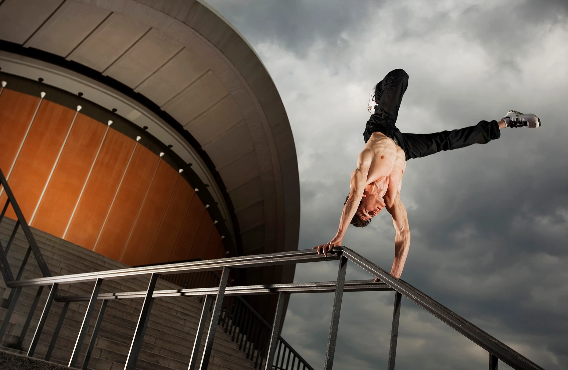 Der Akrobat und Freerunner John Förster posiert mit freiem Oberkörper einen freestyle- Handstand am Geländer vor der schwangeren Auster in Berlin bei bedecktem Himmel. Eine Freie Arbeit.