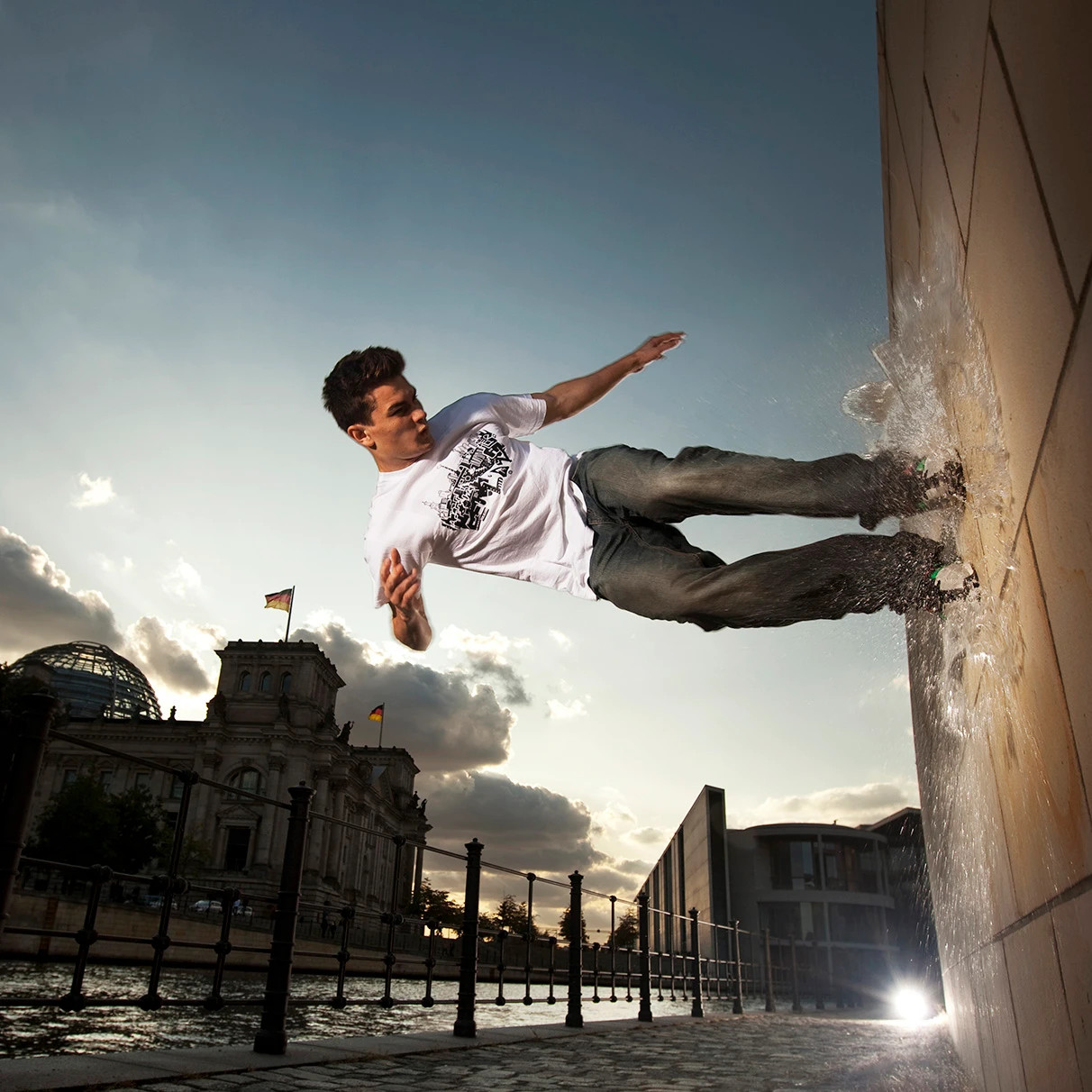 Der Akrobat und Freerunner John Förster springt an eine Steinmauer in eine Pfütze. Dabei handelt es sich um einen analogen Trick. Im Hintergrund die Spree und der Reichstag im Gegenlicht mit freiem Himmel. Eine Freie Arbeit.