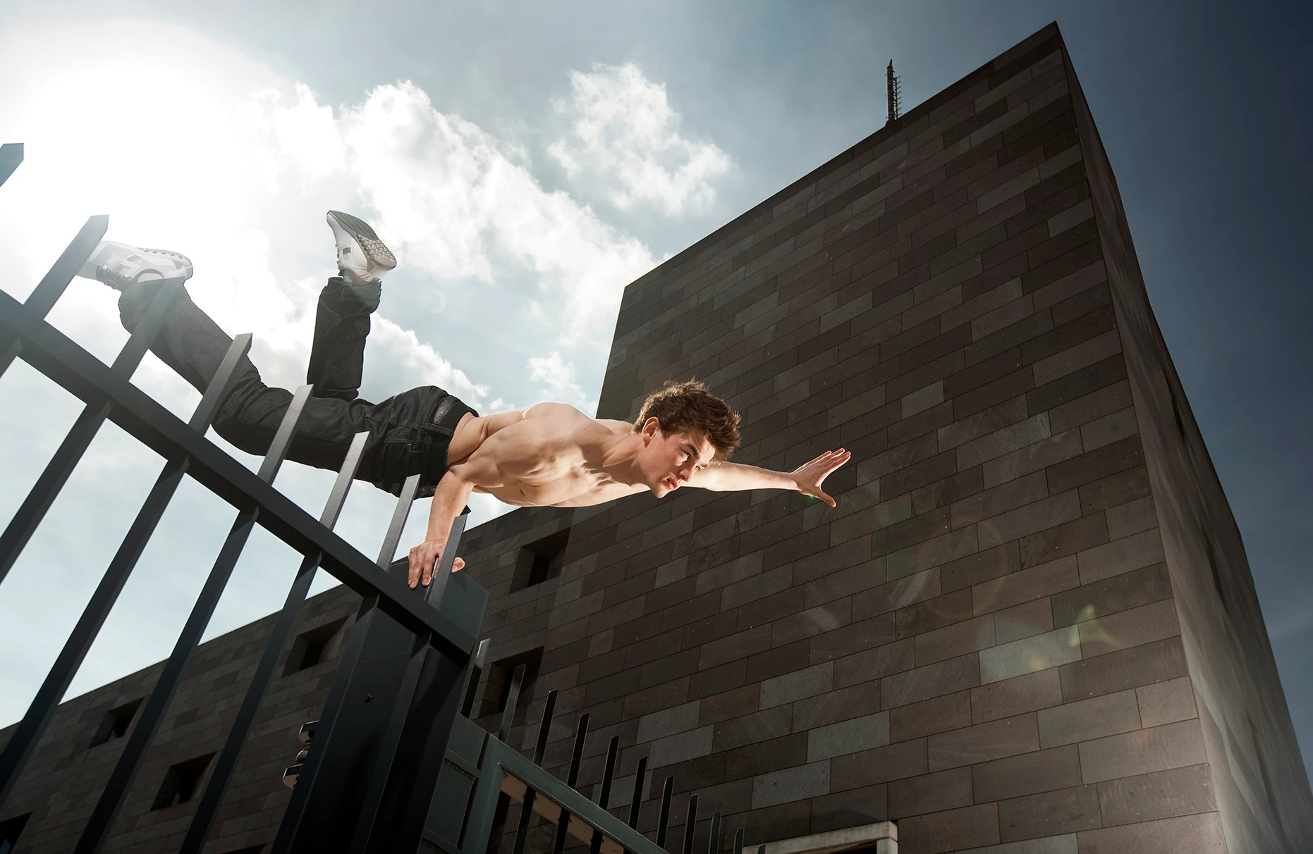 Der Akrobat und Freerunner John Förster posiert mit einer Hand in einer Art Superman- Pose auf einem hohen und massiven Stahlzaun mit Spitzen. Freier Oberkörper. Im Hintergrund ein modernes Gebäude mit Steinfassade. Eine Freie Arbeit.