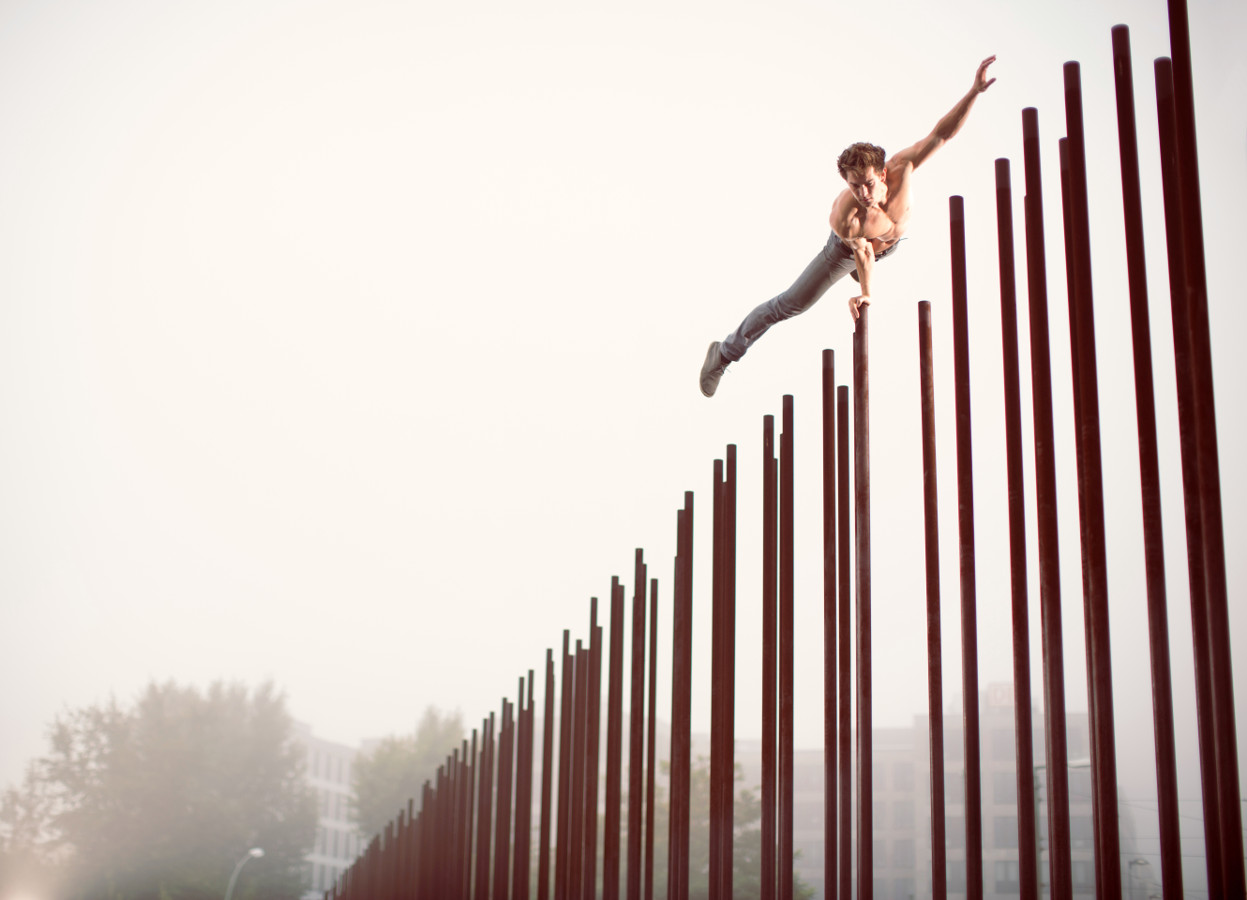John Förster springt dynamisch über das Stelenfeld der Gedenkstätte Berliner Mauer, Bernauer Straße in Berlin, gestreckt im Flug – dynamische Sport- und Konzeptfotografie, 100% real ohne Bildbearbeitung.