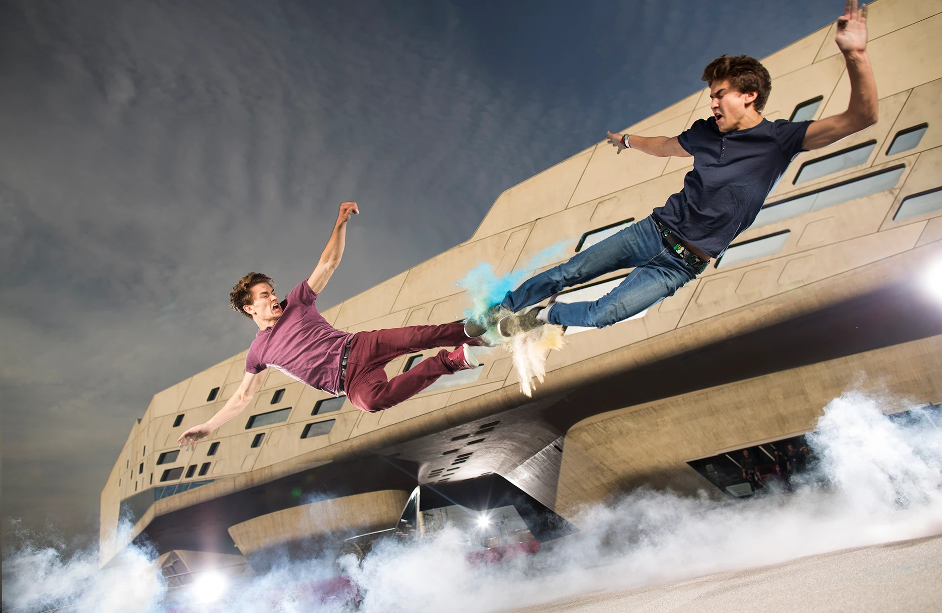 Die Akrobaten und Freerunner John und Jim Förster springen einen high five mit den Füssen. Dabei explodiert farbige Staubwolke zwischen ihren Füssen. Dies ist ein analoger Trick. Viel künstlicher Nebel am Boden und das Phaeno Wolfsburg im Hintergrund. Eine Freie Arbeit.