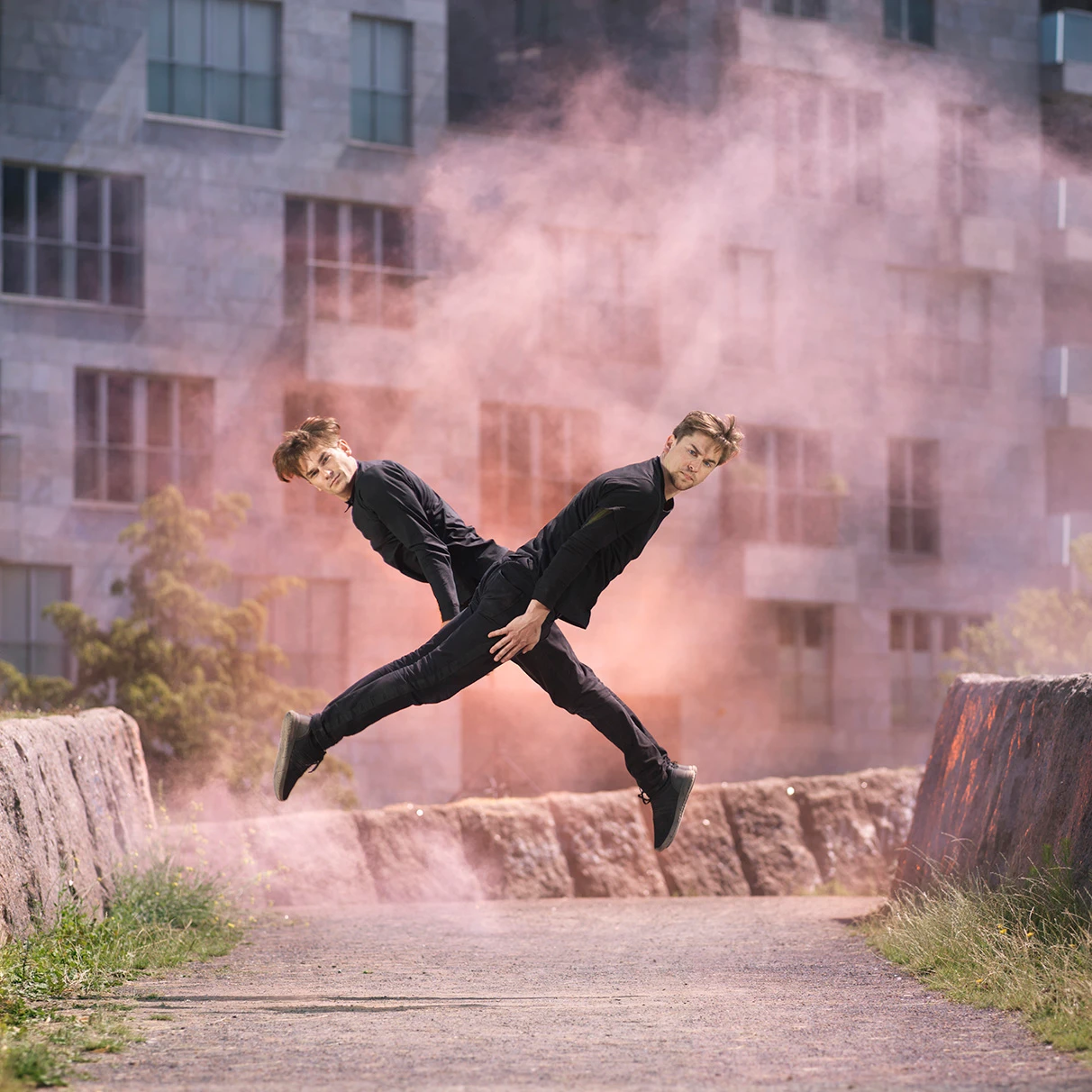 Die Akrobaten und Freerunner John und Jim Förster springen gleichzeitig eine Kreuz- Formation und blicken in die Kamera. Links und rechts werden sie von Granitmauern eingerahmt und hinter ihnen schwebt oranger Nebel, ein analoger Trick. Im Hintergrund moderne Gebäude mit Steinfassade, Potsdamer Platz Berlin. Eine Freie Arbeit.