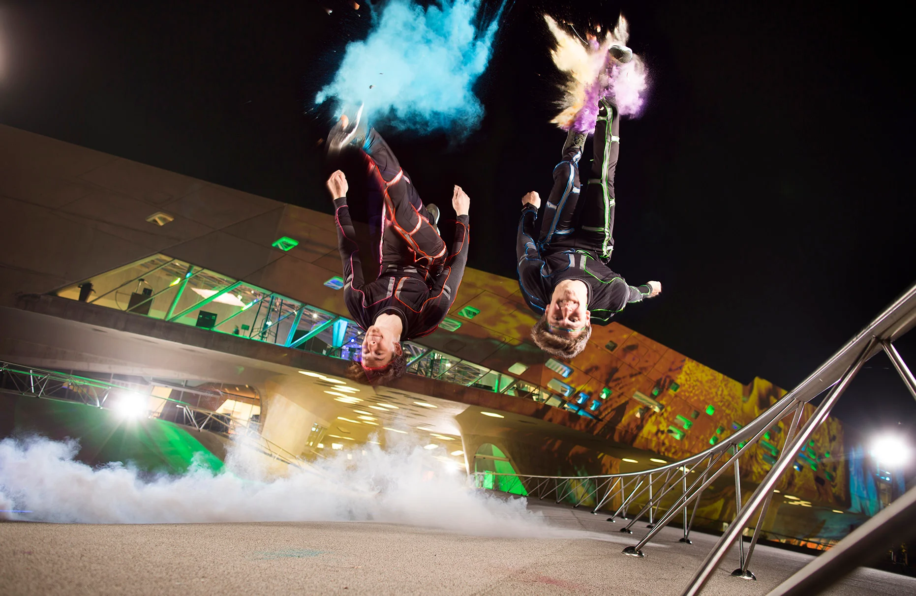Die Akrobaten und Freerunner John und Jim Förster springen gleichzeitig einen Salto. Dabei explodiert farbige Staubwolke an ihren Füssen. Dies ist ein analoger Trick. Viel künstlicher Nebel am Boden und das Phaeno Wolfsburg farbig angetrahlt im Hintergrund bei Nacht. Eine Freie Arbeit.