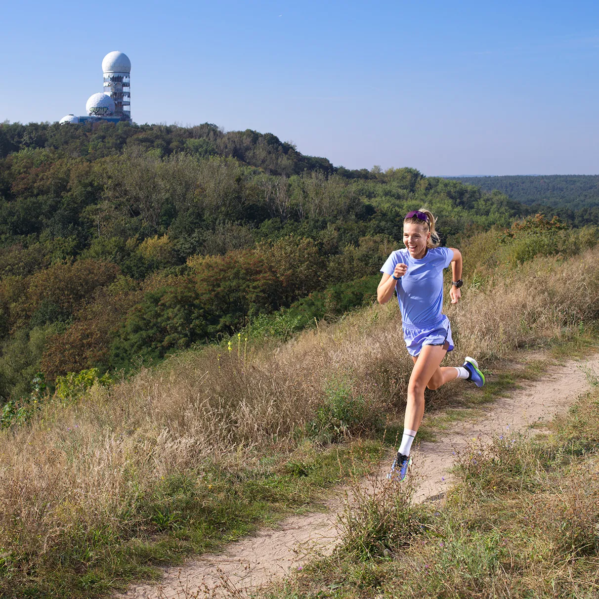 Die Sportlerin und Läuferin Joyce Hübner joggt fröhlich lachend auf dem Drachenberg in Berlin bei herlichem Sonnenschein. Alles ist grün und im Hintergrund ist der Teufelsberg mit der bekannten Radaranlage zu sehen. Produziert für Runners World Magazin mit Hasselbald 501c CFVii50v.