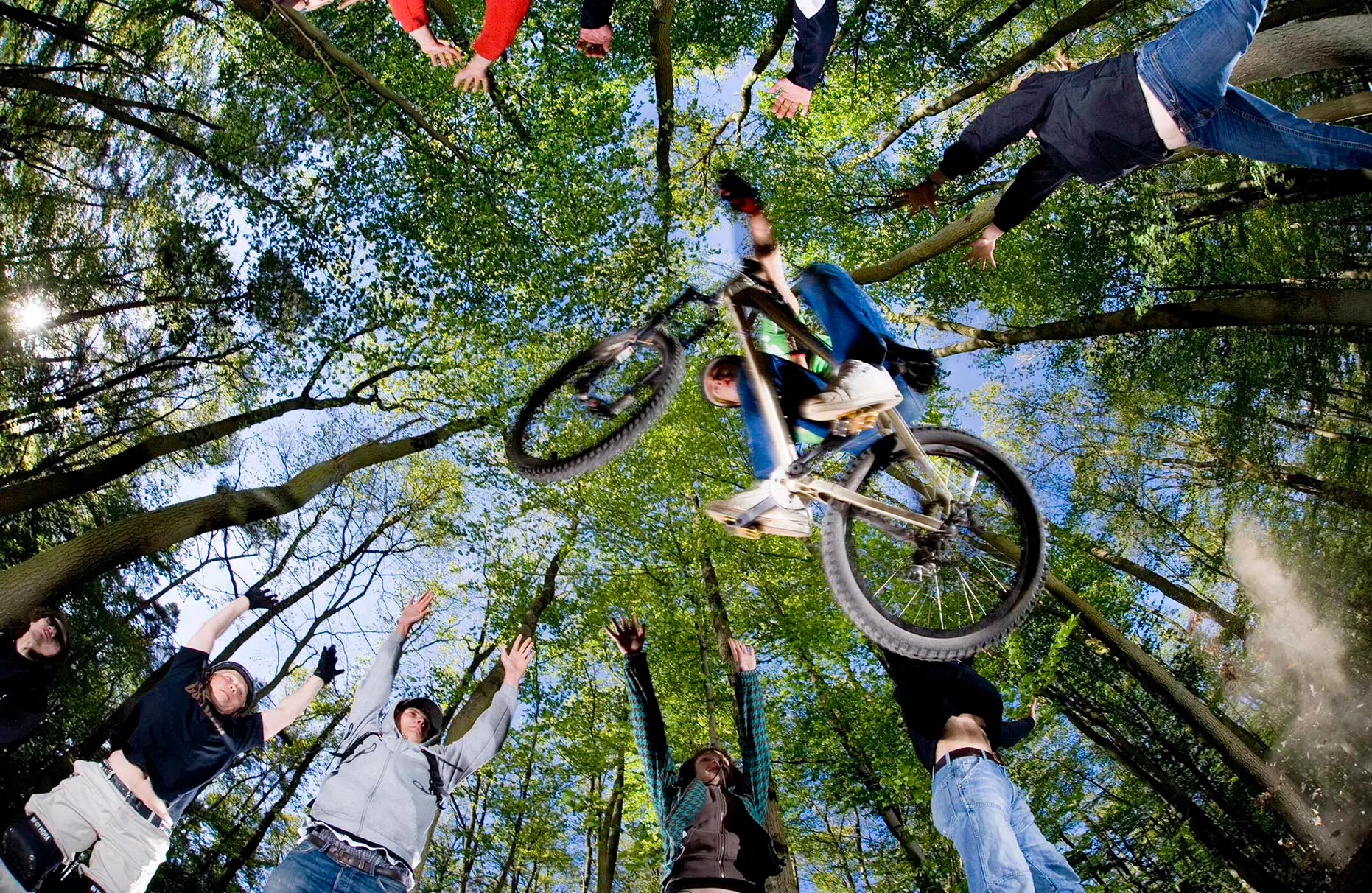 Eine Mountain-Bike- Fahrer springt in einem Laubwald über die am Boden liegende Kamera. Mit ihm springen ringsum Freunde in die Luft. Eine Freie Arbeit.