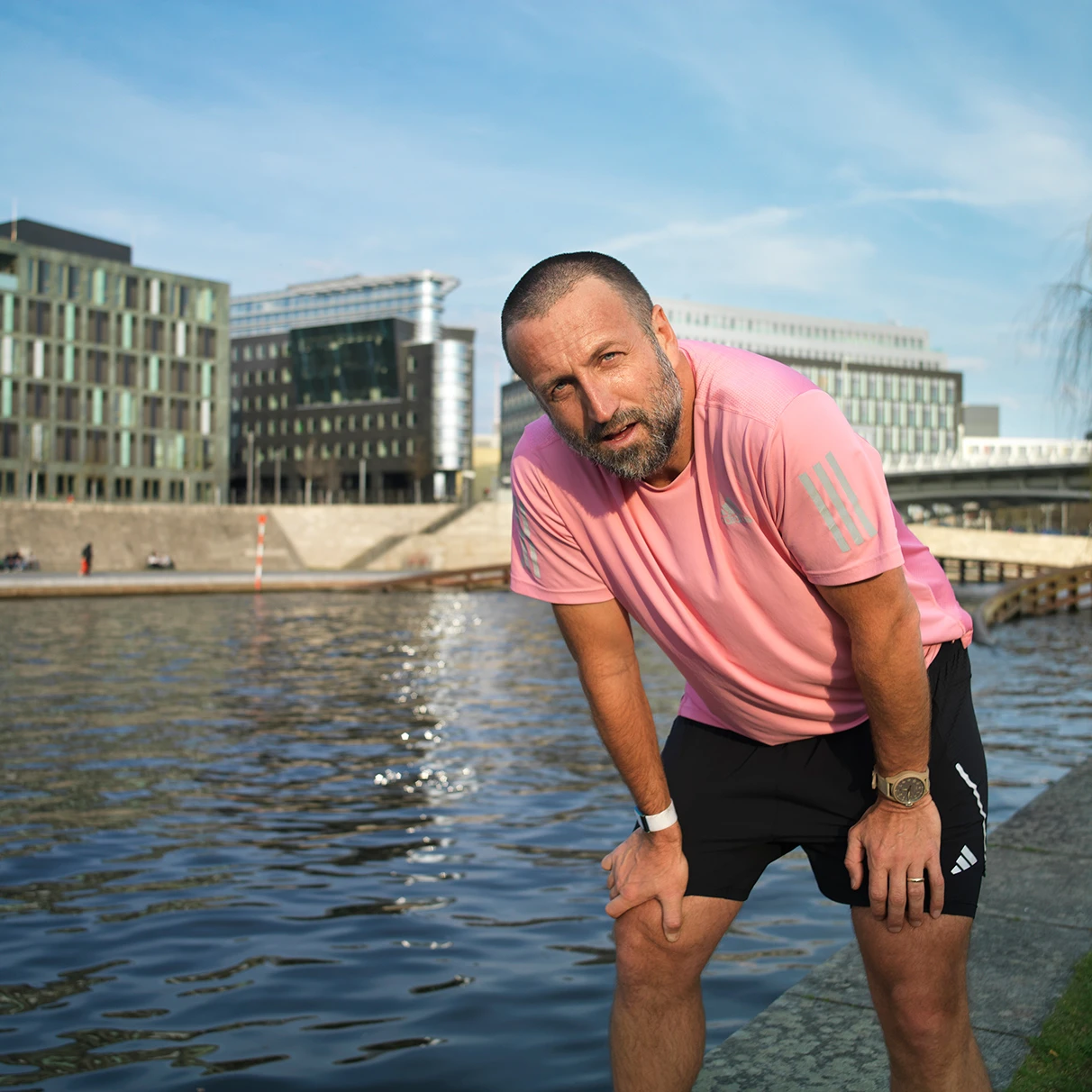 Paul Ripke posiert in einer Verschnaufpausen- Situation bei einem Jogging-Lauf und blickt in die Kamera. Im Hintergrund die Spree und moderne Gebäude mit blauem Himmel. Prodziert für Runners World Magazin mit Hasselblad 501c CFV 16.