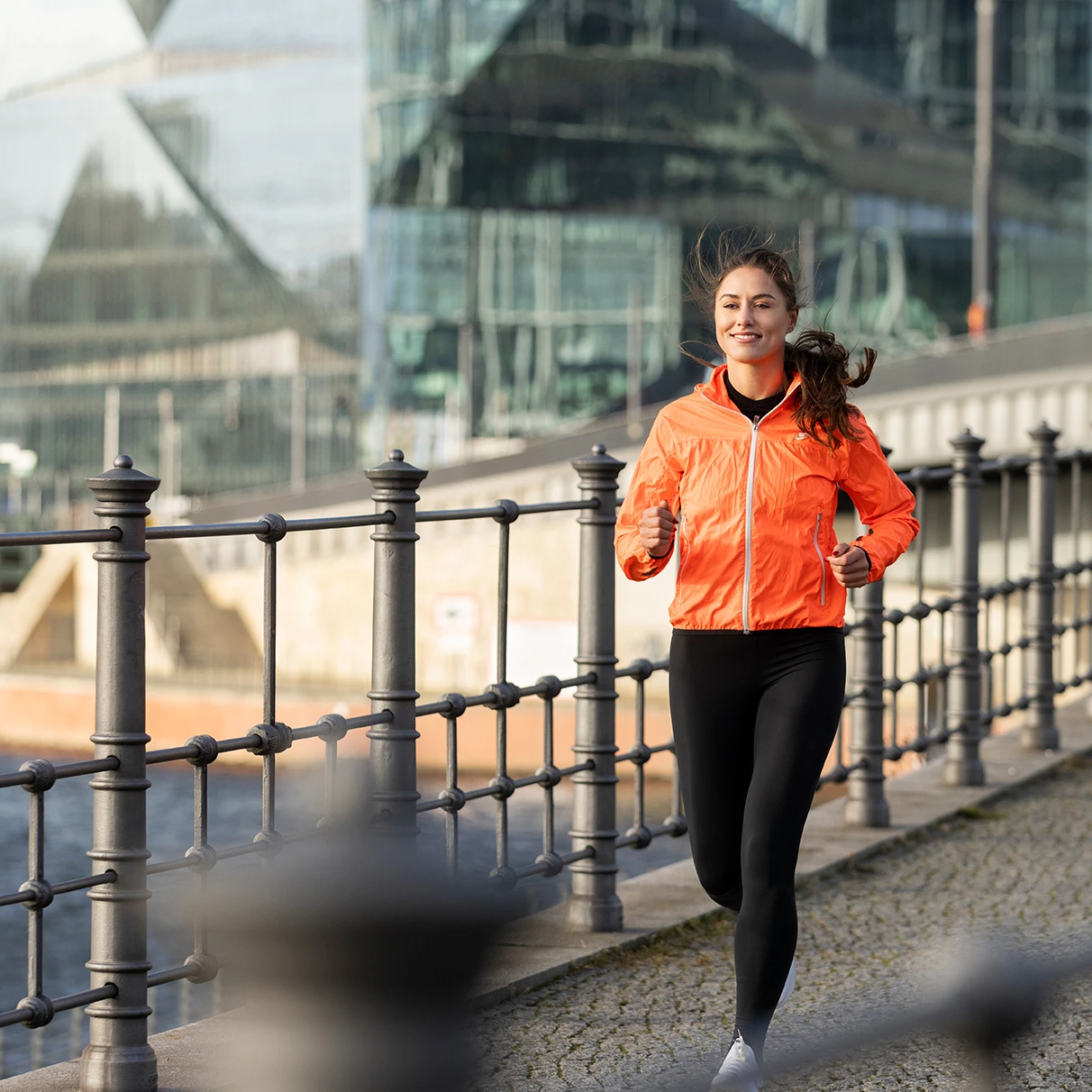 Sherin Maruhn joggt an der Spree an einem Geländer vorbei und lacht fröhlich. Im Hintergrund ein formatfüllendes modernes Glasgebäude. Nähe Hauptbahnhof. Produziert für Runners World Magazin.