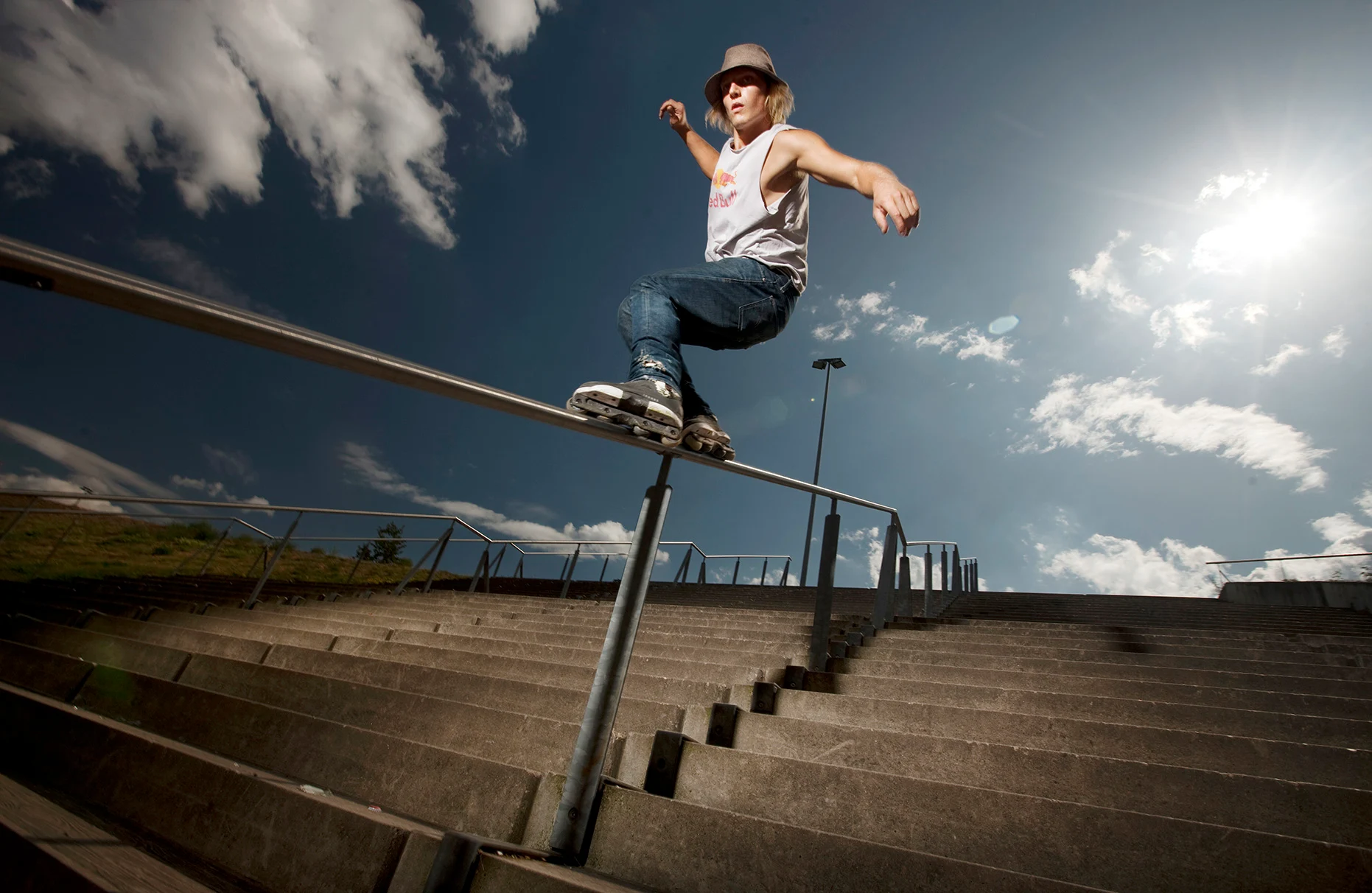 Der inline-skater Stefan Horngacher grindet mit seinen inline skate's über ein Edelstahl- Geländer am Berliner Velodrom. Im Hintergrund nur Betontreppen und blauer Himmel mit weissen Wolken. Produziert für Red Bull.