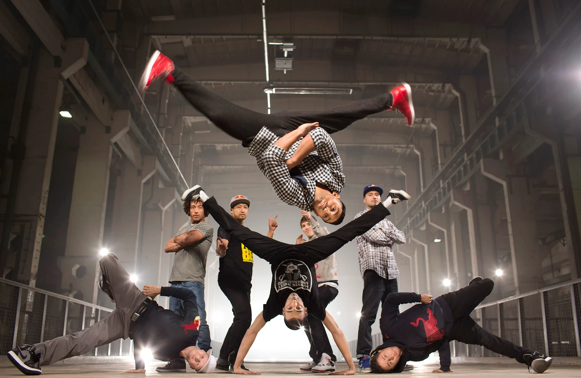 Acht Mitglieder der berühmten breakdance crew Flying Steps aus Berlin posieren in einer ausgeleuchteten Industriehalle für die Ankündigung ihrer Tour Flying Bach. Einer von ihnen springt einen Salto im Spagat. Produziert für Red Bull.