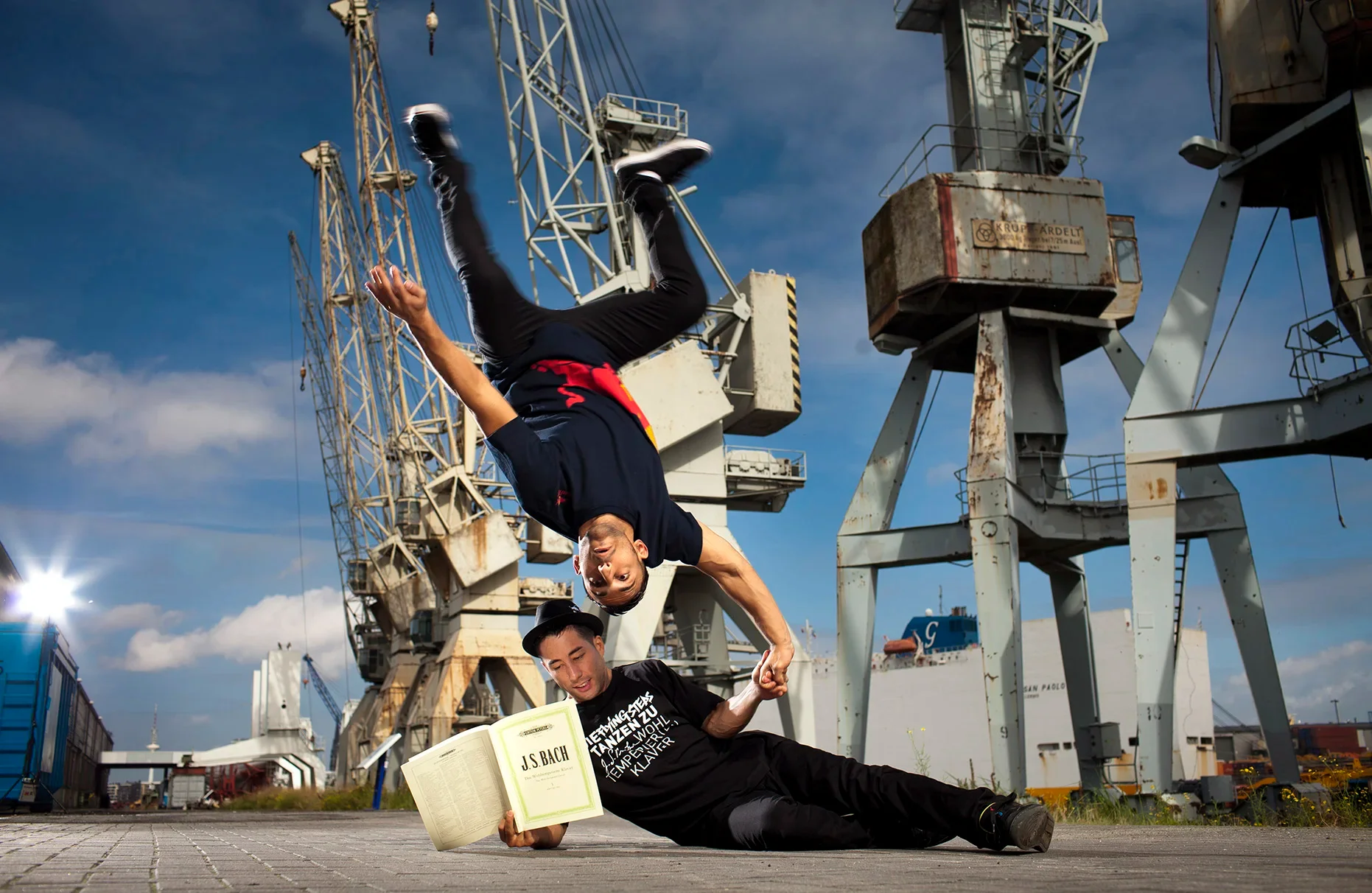 In einer Hamburger Hafen-Kulisse mit historischen Kränen und blauem Himmel posieren die breakdancer Benny Kymoto und Gengis Ademoski von den berühmten Flying Steps für die Ankündigung ihrer Tour Flying Bach. Benny Kymoto sitzt auf dem Boden und liest in einem Bach Notenbuch, während Gengis Ademoski kopfüber mit seiner Hand auf Bennys Hand steht und auch ins Buch schaut . Produziert für Red Bull.