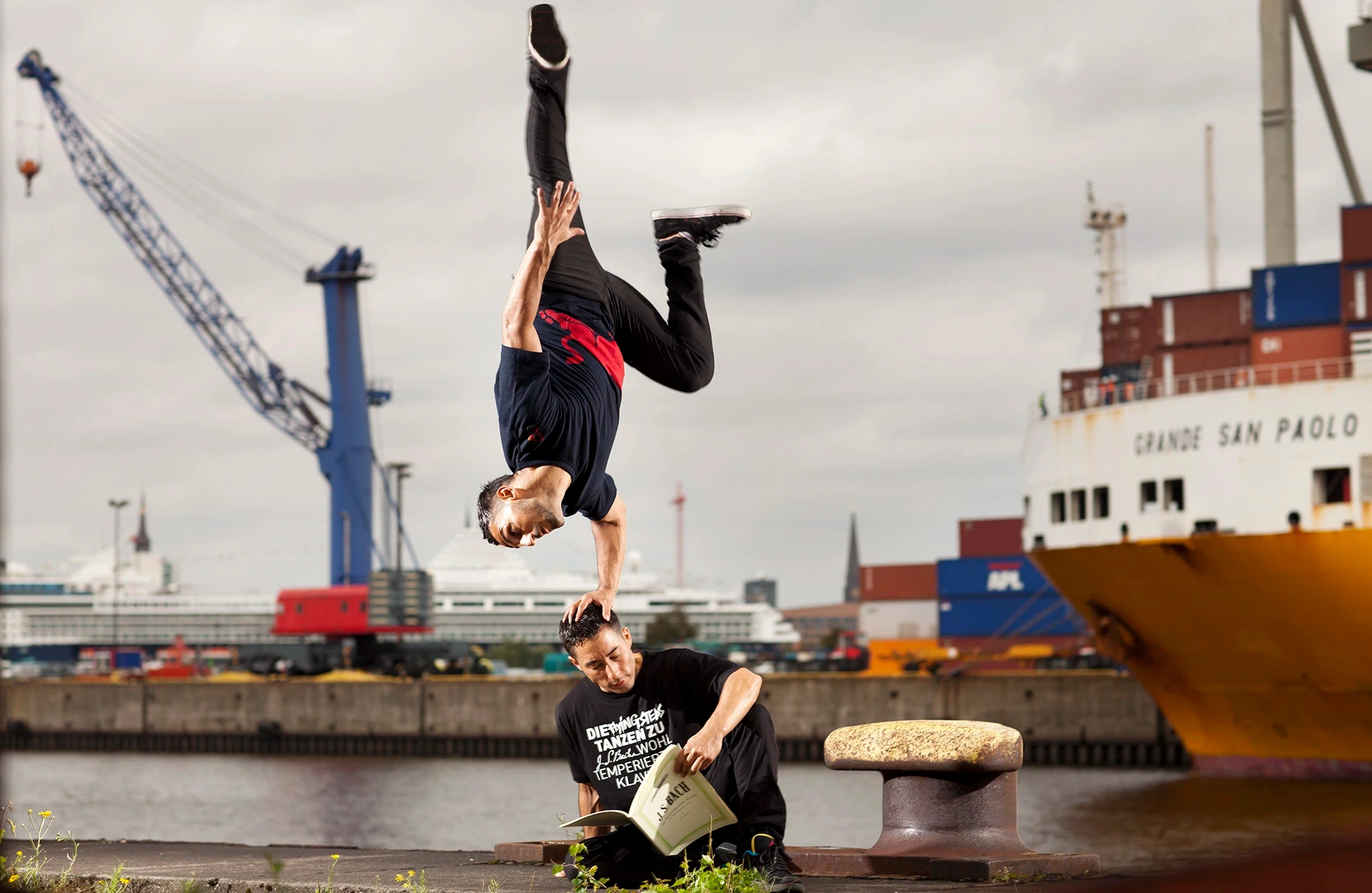 In einer Hamburger Hafen-Kulisse mit Kran und Frachtschiff posieren an der Hafenkante die breakdancer Benny Kymoto und Gengis Ademoski von den berühmten Flying Steps für die Ankündigung ihrer Tour Flying Bach. Benny Kymoto sitzt auf dem Boden und liest in einem Bach Notenbuch, während Gengis Ademoski kopfüber mit seiner Hand auf Bennys Kopf steht und auch ins Buch schaut . Produziert für Red Bull.