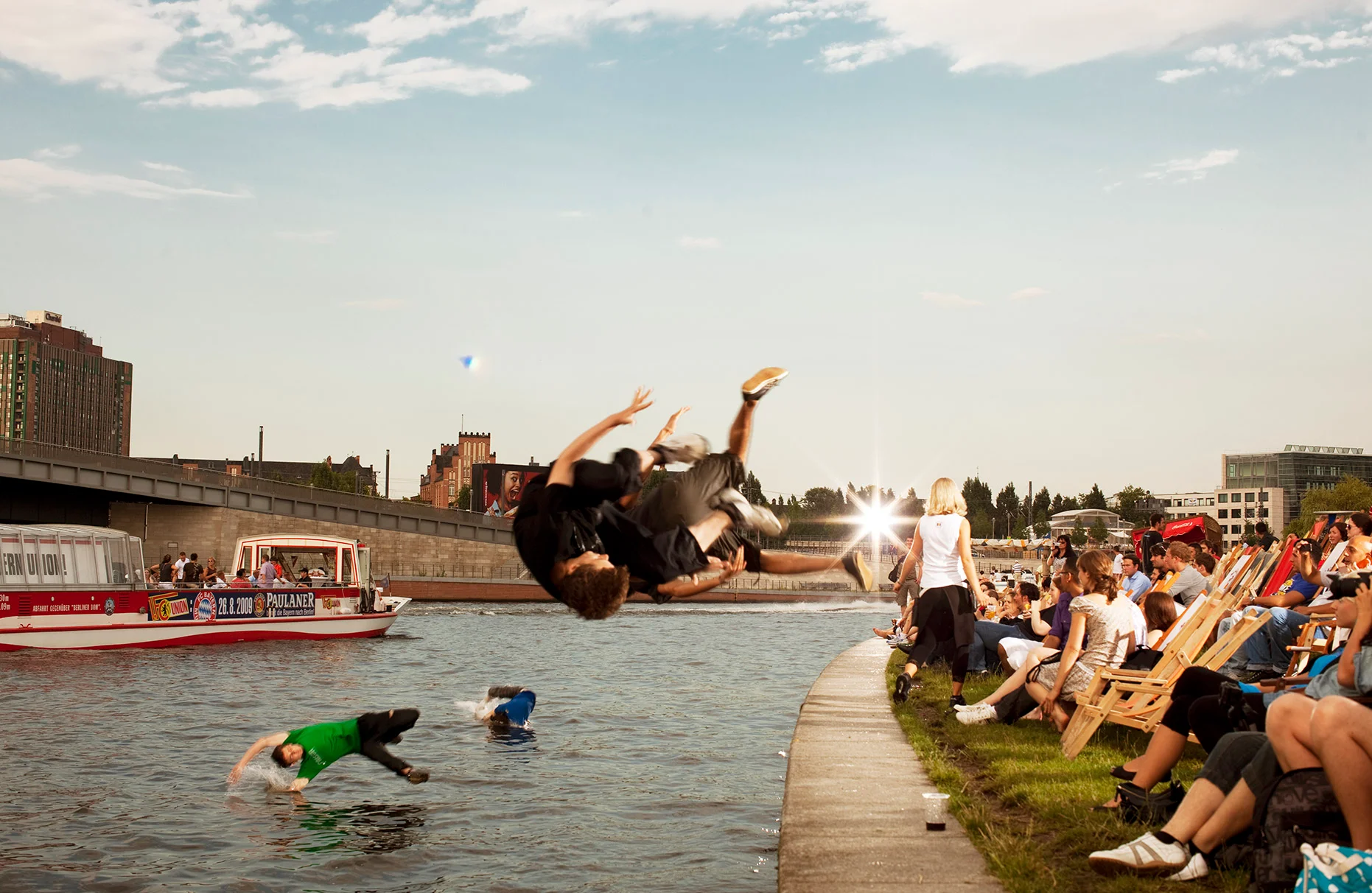 Mehrere junge freerunner Sportler springen gleichzeitig an einem sonnigen Tag mit Salto in die urbane Spree. Ein Fahrgastschiff fährt vorbei. An der Uferkante sitzen auf Liegestühlen viele Strandbar- Gäste und staunen, denn die Aktion geschah bis zur letzten Sekunde verdeckt. Produziert für das Rausch- Magazin zum Thema Mutprobe.