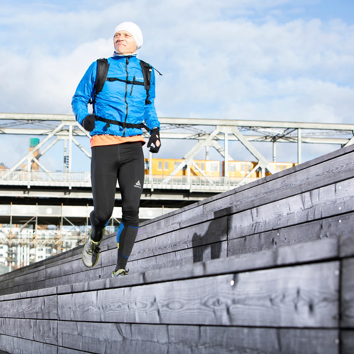 Der Läufer Mark Brunner joggt in blauer Winter- Sportbekleidung auf einer Art Tribüne aus Holz im Gleisdreieck- Park in Berlin. Im Hintergrund eiserne S-Bahn- Brücke und blauer Himmel mit Wolken. Produziert für Runners World Magazin.